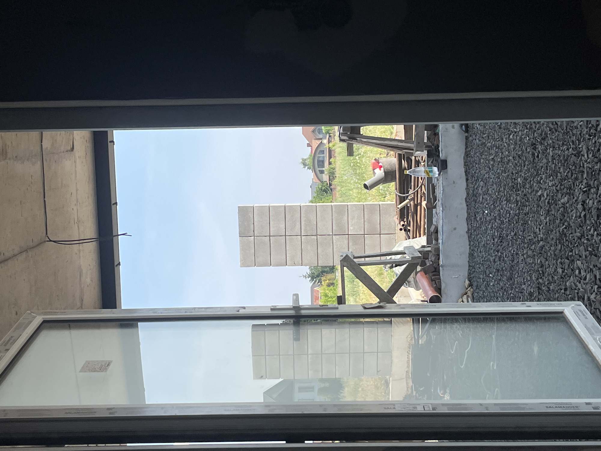 The image captures a view through an open doorway onto a construction site in Strumivka, Ukraine. In the foreground, a partially open white-framed window with clear glass reflects the sky and the surrounding area. The construction site itself features stacks of gray concrete blocks, one of which is a tall, narrow column. Adjacent to this is a wooden structure with diagonal supports. Several pieces of construction material, including pipes and a red-orange pipe, are visible. A bucket containing some red items and a gray pipe is also present. A plastic water bottle sits on a ledge near the bucket. In the background, beyond the immediate construction zone, several houses with pitched roofs and trees are visible under a clear blue sky. The ground in the foreground and mid-ground is covered with gravel. The overall scene depicts an active or recently active construction area.