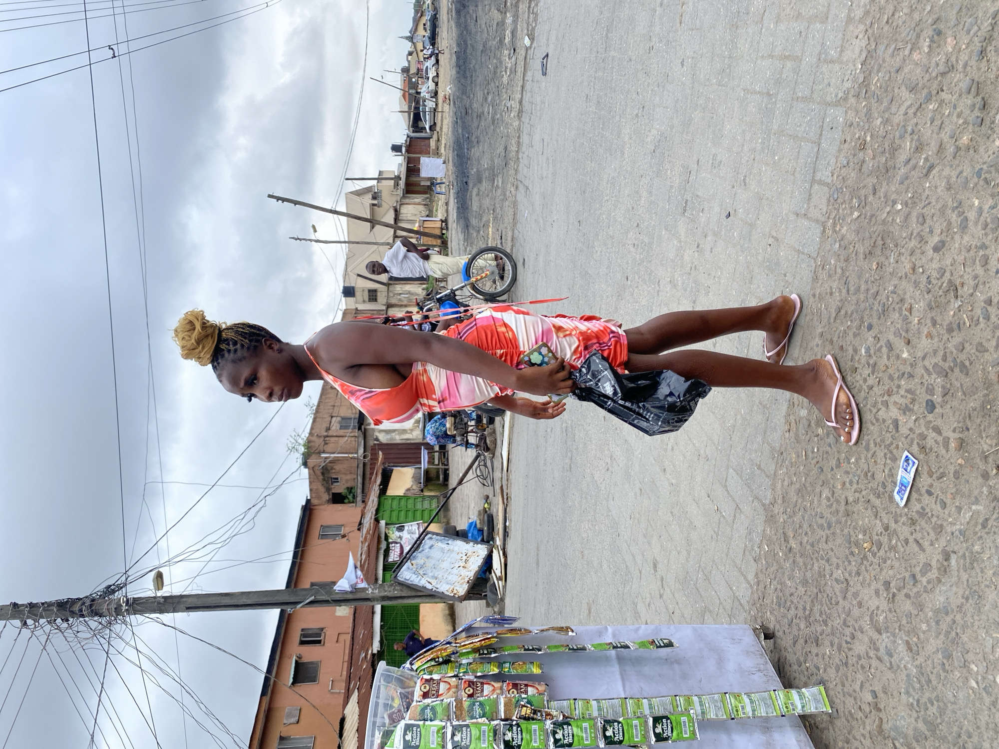 In an outdoor urban setting, a young woman with blonde braided hair styled in a bun stands to the right, facing left and looking away from the camera. She wears a vibrant orange, pink, and white patterned dress that is off-the-shoulder and features a tie-back detail. In her right hand, she holds a black plastic bag. Her left hand rests on the bag. She is wearing pink flip-flops. A condom wrapper is visible on the ground in front of her.
To the left of the woman, a white display stand is laden with various packaged goods, predominantly green and white packages labeled "Action Bitters." Further to the left, a utility pole is visible, heavily entangled with a complex network of electrical wires.
In the background, buildings with orange and brown facades line the street. A blue motorcycle with chrome accents is parked behind the woman. A man in a white shirt and light-colored pants stands behind the motorcycle, looking towards the camera. The ground is paved with concrete and stones. The sky is overcast with clouds. The overall scene suggests a street-level market or commercial area in an urban environment.