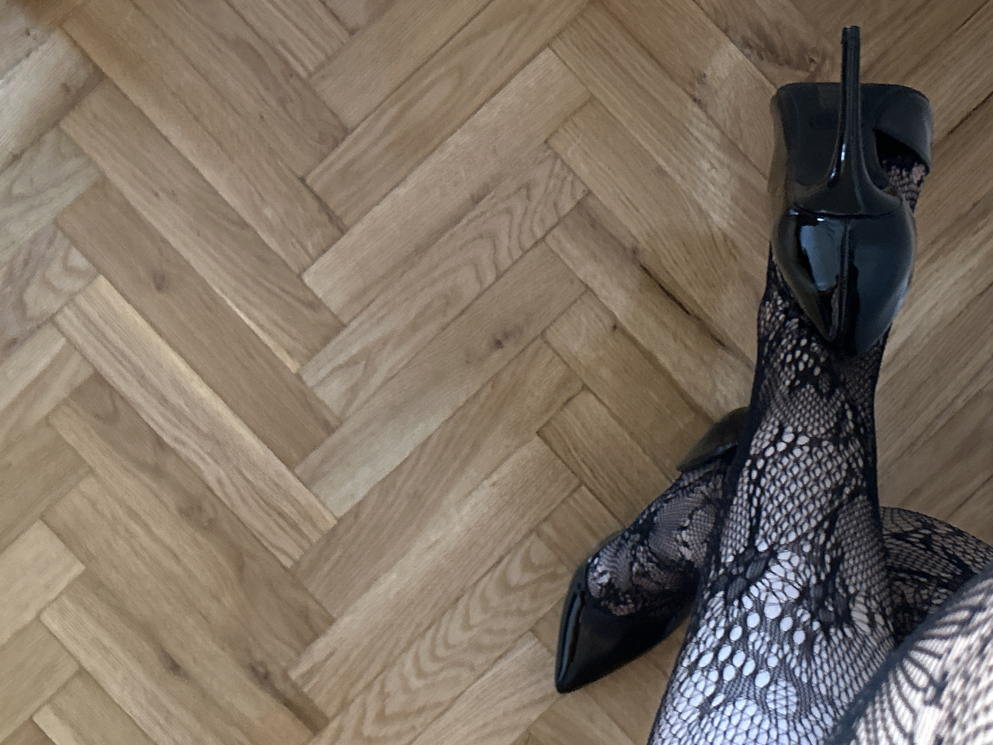 A pair of legs wearing black patterned stockings and black patent leather high heels are positioned on a light-colored herringbone wood floor. The legs are crossed. The location cannot be determined to be in Novi Sad, Serbia.