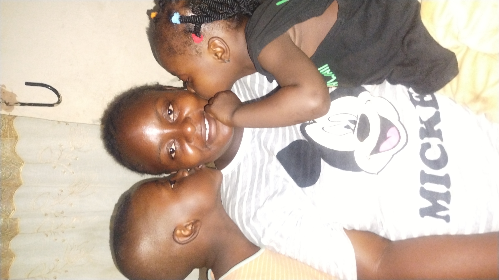 The image captures three individuals: an adult female positioned centrally, flanked by two young children. The adult female, with dark skin and short dark hair, smiles slightly, revealing teeth. She wears a light-colored, horizontally striped t-shirt featuring a large black and white Mickey Mouse graphic on the front, with the word "MICKEY" printed below.
To the adult's left, a young boy with dark skin and a closely cropped haircut leans in, kissing her left cheek. He wears a light orange or striped sleeveless top. To the adult's right, a young girl with dark skin and dark braided hair adorned with colorful beads (red, blue, yellow) is simultaneously kissing her right cheek. She wears a black sleeveless top with partially visible green text on the front.
The background indicates an indoor setting, likely in the city of Jos, Nigeria. It features a light-colored wall on the right, with a dark, U-shaped hook attached to it in the upper right quadrant. To the left, a light-colored curtain with a decorative gold-patterned valance at the top is visible. A yellowish, textured fabric is present in the lower right foreground. The overall interaction conveys affection between the individuals.