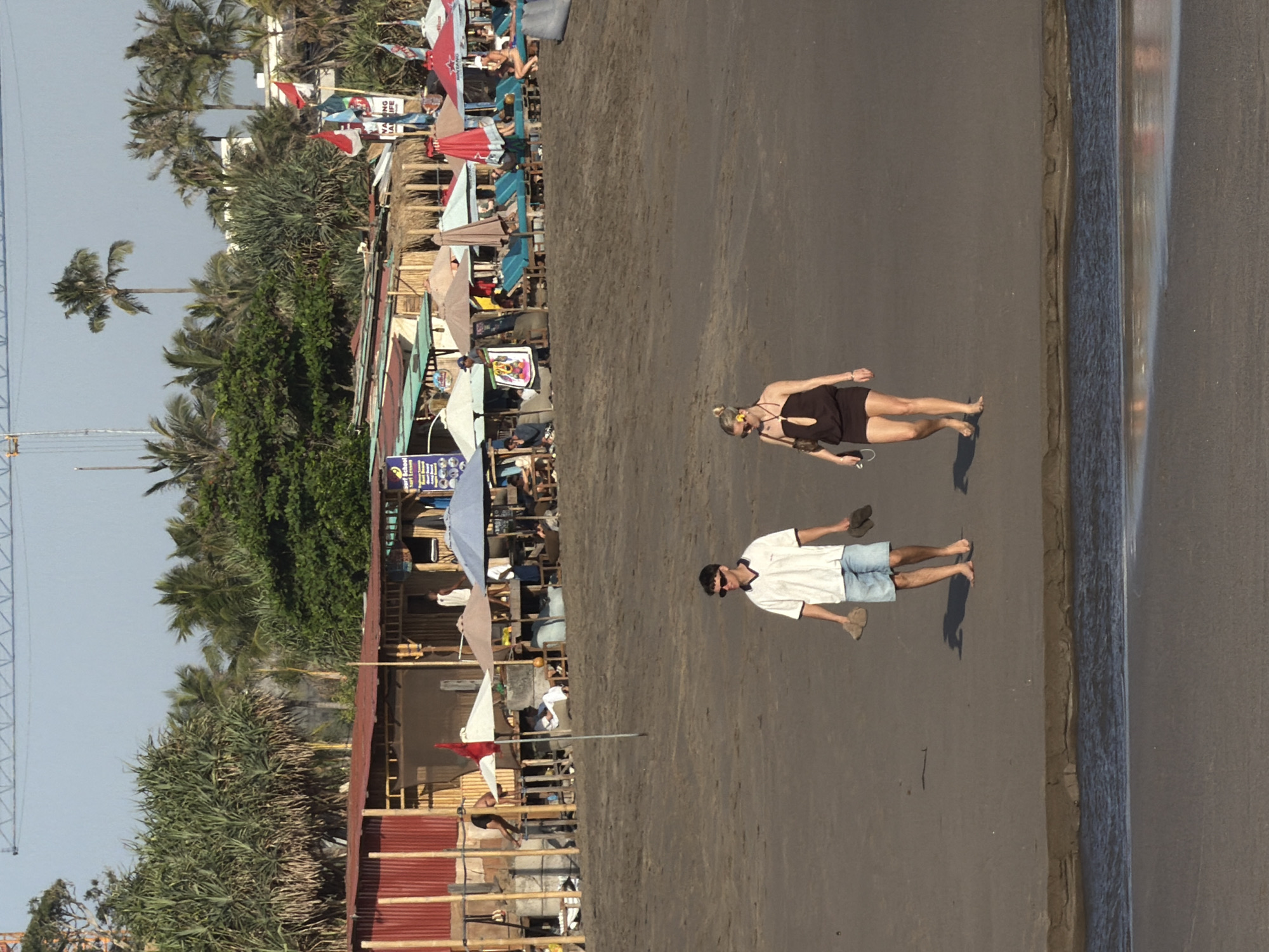 The image depicts a beach scene in North Kuta, Indonesia, at dusk. In the foreground, a man and a woman walk together on the sandy shore. The man, wearing a white polo shirt and blue shorts, carries a pair of flip-flops in each hand. The woman, in a dark strapless dress and sunglasses, also walks barefoot. Behind them, a row of beach umbrellas and lounge chairs are set up, with some people relaxing. A low-rise building with a red roof and bamboo structures lines the back of the beach. Palm trees and lush vegetation are visible behind the buildings. The sky is clear with a hint of orange, suggesting the sun is setting. A crane structure is visible in the far distance, indicating ongoing construction or development.