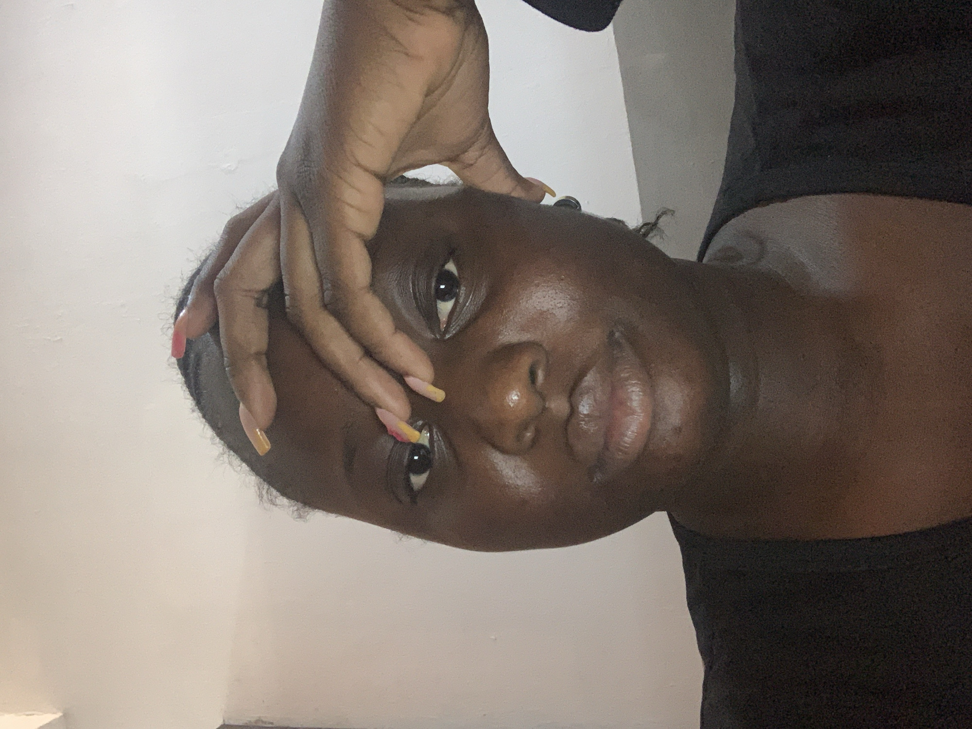 A close-up, front-facing portrait shows a young Black woman indoors, positioned in the center of the frame. She is looking directly at the camera with her left hand resting on her forehead, obscuring part of her brow and eye. Her fingernails are long and painted with a gradient of yellow and pink. Her lips are slightly parted, and her eyes are dark and open. She is wearing a black v-neck top. The background consists of a plain, light-colored wall with some texture and a subtle shadow. The lighting is soft, coming from the front and slightly above, creating a subtle sheen on her skin.