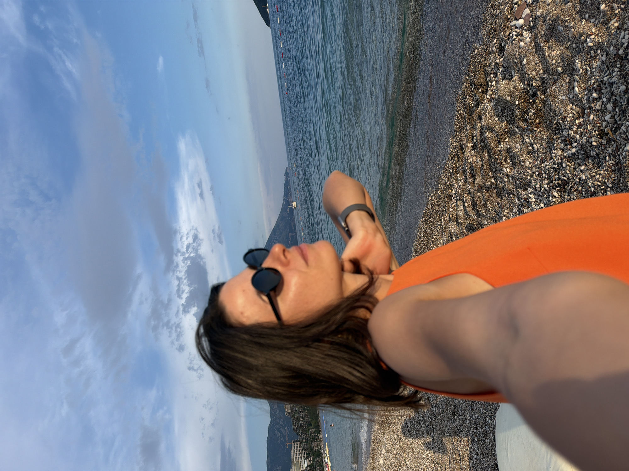 In Budva, Montenegro, a woman with dark hair and sunglasses is seen from the chest up, facing right. She wears an orange tank top and her right arm is extended, holding a phone to take a selfie. Her left hand is on the back of her neck. The background shows a pebble beach leading to the Adriatic Sea, with buoys marking an area in the water. In the distance, buildings are visible on a hillside, and mountains are on the horizon under a cloudy blue sky. The scene is illuminated by natural light, suggesting it is daytime.
