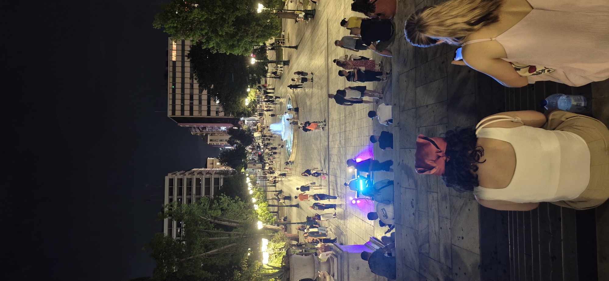 This is a vertical, eye-level nighttime photograph taken in Kotzia Square, Athens, Greece. The image captures a bustling public square illuminated by streetlights, building lights, and colored stage lighting from a temporary setup on the paved ground. In the foreground, two women are seated on broad stone steps, facing away from the camera, looking towards the square. The woman on the left wears a white tank top and tan pants, with a red bandana tied around her head. The woman on the right wears a light pink dress and is looking at her phone.
The middle ground of the image shows a wide, paved plaza filled with people walking and gathering. A circular fountain with illuminated water jets is a central feature in the mid-distance. To the left of the fountain, a small stage is set up with performers and lighting equipment, drawing a crowd. People of various ages and attire are seen milling about, some seated on benches and others standing.
In the background, the scene is framed by buildings on either side of the square, with trees lining the perimeter. The architecture appears to be typical of urban Athens, with multi-story buildings and illuminated windows. The overall atmosphere is lively and social, indicating a public event or a popular gathering spot at night.