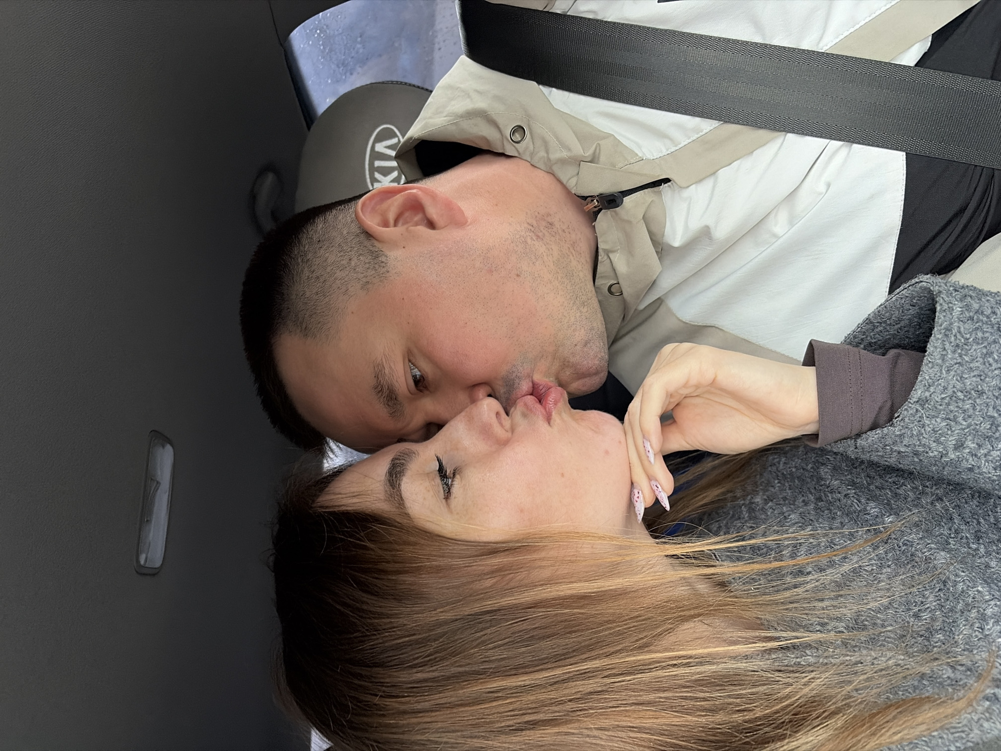 A couple is kissing in a car. The man has short, dark hair and is wearing a light-colored jacket. The woman has long, light brown hair and is wearing a gray sweater. They are both wearing seatbelts. A "KIA" logo is visible on the headrest of the seat. The car's interior is visible in the background. The location is not specified.