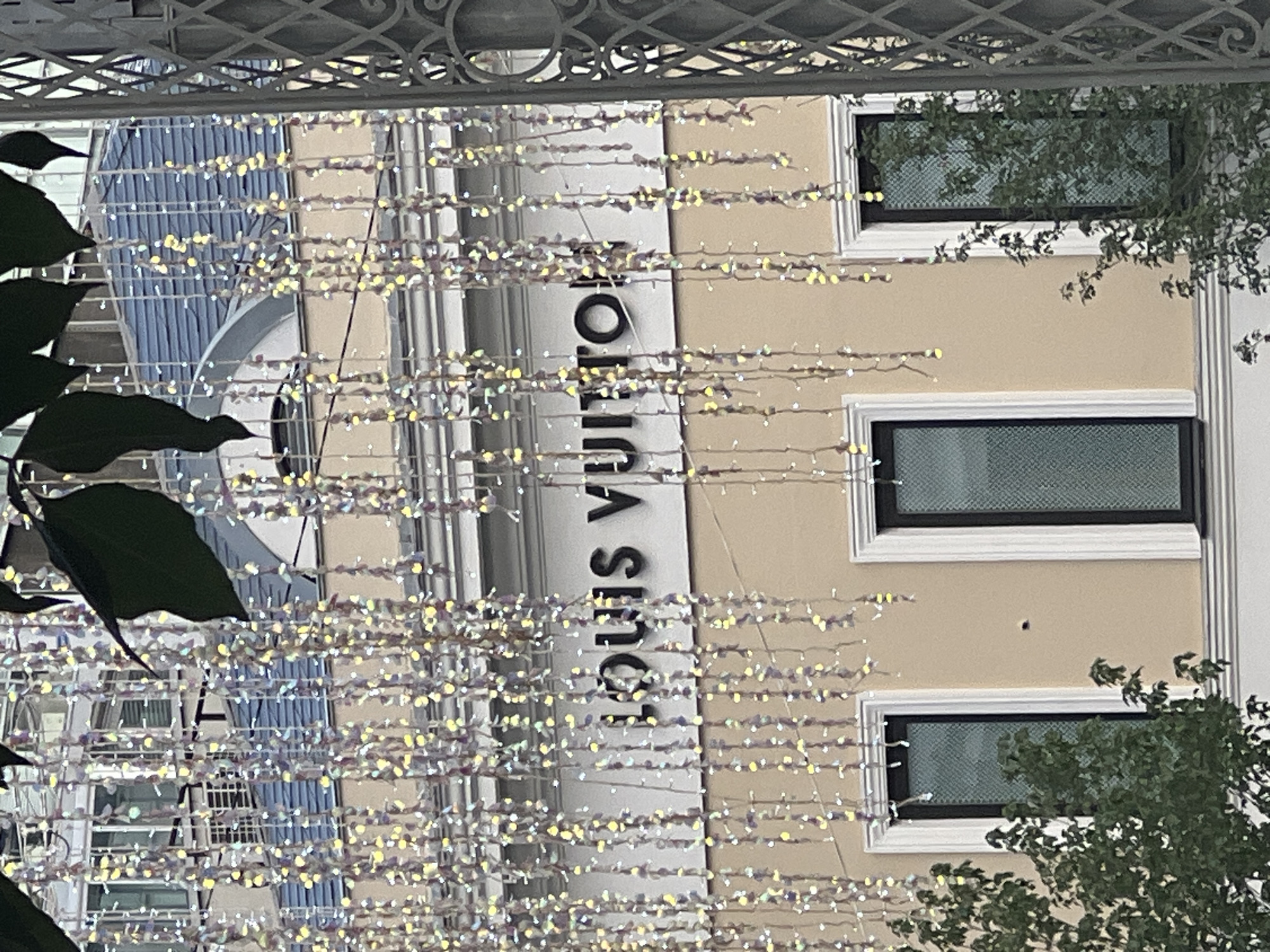 The image shows a building with the "LOUIS VUITTON" logo prominently displayed on its facade. The building is light beige and has several windows with dark frames and textured glass. The Louis Vuitton sign is in large, dark, capitalized letters vertically oriented. In front of the building, there are strings of small, sparkling lights, some of which appear to be in shades of yellow and purple. In the foreground, dark green leaves are visible, partially obscuring the view of the lights and the building. The leaves suggest that the photo may have been taken outdoors, possibly from a park or garden area. The presence of the Louis Vuitton store indicates an urban or high-end commercial setting. The lighting suggests it could be dusk or evening, as the string lights are illuminated. The image does not clearly depict any people or specific activities, focusing more on the architectural elements and the branding. There are no obvious weather cues, but the lighting implies artificial illumination is active. The backdrop appears to be a building with a blue-toned facade, possibly reflecting the sky or other structures. The overall impression is one of a commercial establishment, likely a luxury brand store, adorned with decorative lighting.