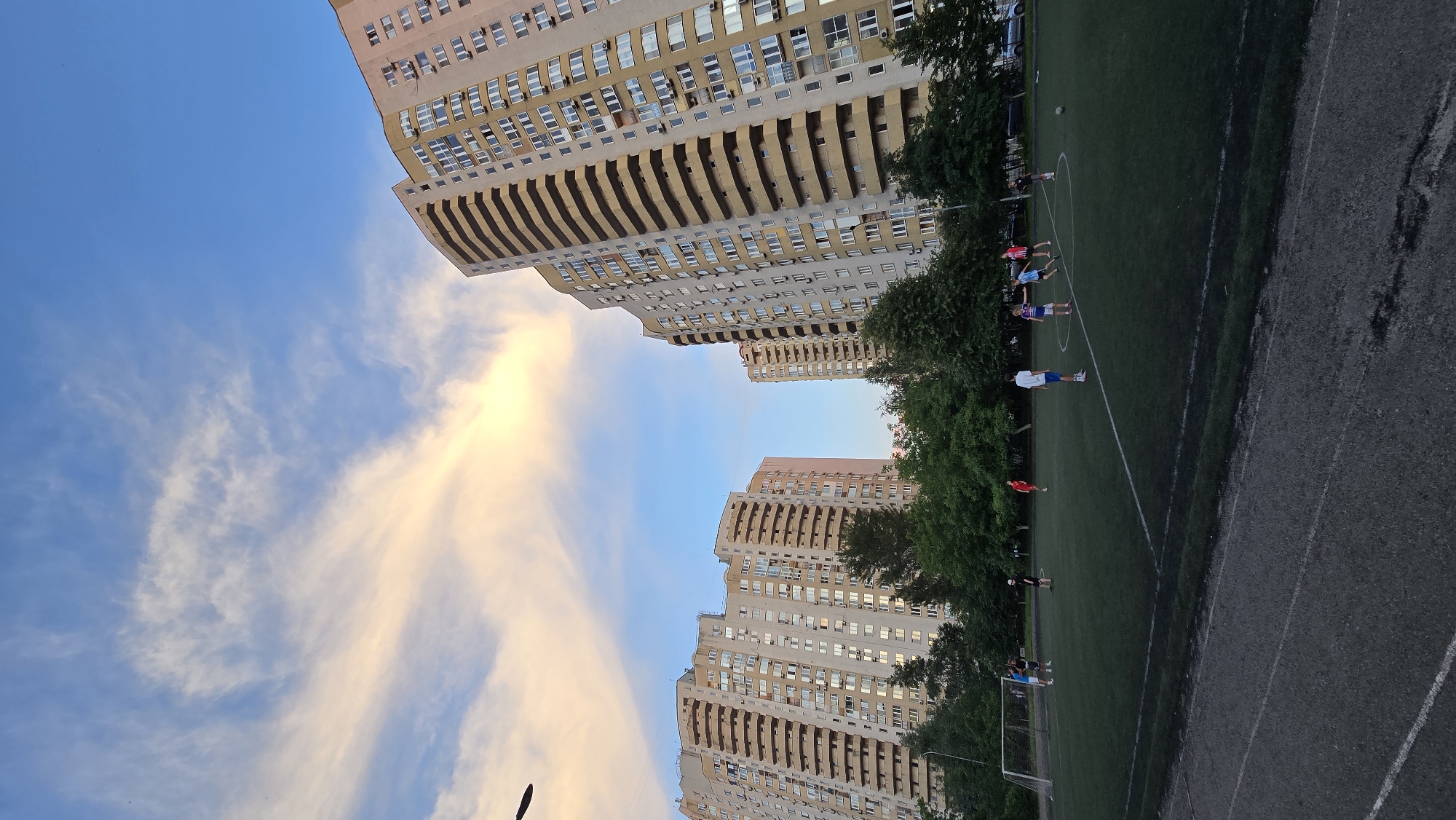 The image depicts a bright, overcast sky with streaks of pink and orange clouds. Two large, modern apartment buildings dominate the left and center of the frame, their light-colored facades punctuated by numerous windows and balconies. The buildings are tall and appear to be in an urban or suburban setting.
To the right of the apartment buildings, a green football (soccer) field is visible. Several people, appearing to be young men and boys, are scattered across the field, some in athletic wear, engaged in what looks like a casual game of football. One person is near the goalpost on the left side of the field. Trees line the edge of the field, adding a natural element to the urban landscape. The ground beside the field is a mixture of asphalt and gravel.
The overall scene suggests an afternoon or early evening in a residential area where a local football game is taking place. The lighting and the colors of the sky indicate it might be sunset or sunrise, but the bright, diffused light suggests it is likely overcast. The presence of the apartment buildings and the football field provides context for a community space within a developed area. No visible text is present in the image.