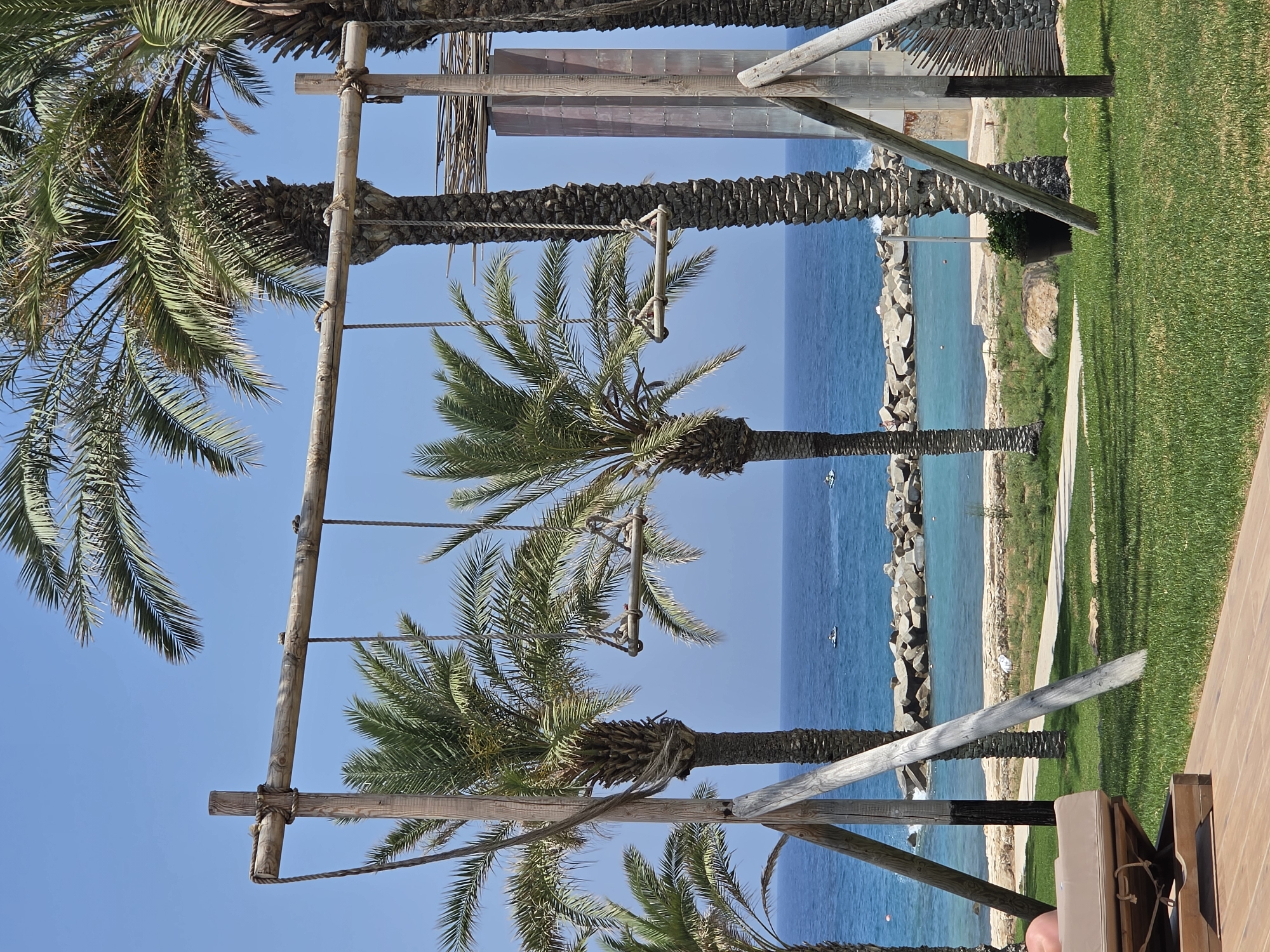 In Hlorakas Community, Cyprus, a tropical scene unfolds with a focus on a handmade wooden swing set, prominently featured in the foreground. The swing set, constructed from rough-hewn timber, supports two individual swings. Each swing consists of a wooden plank suspended by ropes. Surrounding the swing set are several tall palm trees, their fronds casting dappled shadows on the manicured green lawn.
Beyond the lawn, the cerulean expanse of the Mediterranean Sea stretches towards the horizon, dotted with two small white boats. A breakwater made of large grey rocks separates the sea from a tranquil turquoise bay. The shore is lined with sand and sparse vegetation. In the foreground, a portion of a wooden deck and a reclined lounge chair are visible, suggesting a resort or recreational area. The sky is a clear, bright blue, indicative of a sunny day. The overall ambiance is one of serene coastal relaxation.