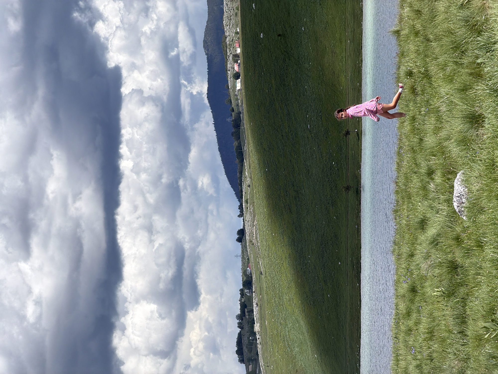 This image captures a wide outdoor landscape in Novakovići, Montenegro, under a sky dominated by heavy, dark grey and white cumulus clouds. The upper two-thirds of the frame are filled with these clouds, featuring a prominent dark grey band horizontally across the mid-sky.
In the mid-ground, a serene body of water, likely a lake, stretches horizontally across the scene with a light blue-green surface. Beyond the lake, rolling green hills ascend, exhibiting distinct patches of sunlight and shadow, suggesting a dynamic interplay of light filtering through the clouds. Scattered small white and red-roofed buildings are visible on these hills, along with sparse trees and what appear to be power lines or utility structures further back. In the distant background, darker, more mountainous terrain rises, covered in denser vegetation.
The foreground consists of lush green grass, interspersed with small purple wildflowers, leading up to the edge of the lake. A small, light-colored rock is situated in the lower-left corner of the foreground. A young girl, approximately 7-10 years old, is depicted in the lower-right quadrant of the image, walking along the grassy bank beside the lake. She is wearing a long-sleeved pink top and pink shorts, with white socks and light-colored shoes. Her body is angled towards the right side of the frame, but her head is turned back over her left shoulder, looking towards the viewer. Her posture indicates mid-stride, with her left leg advanced.