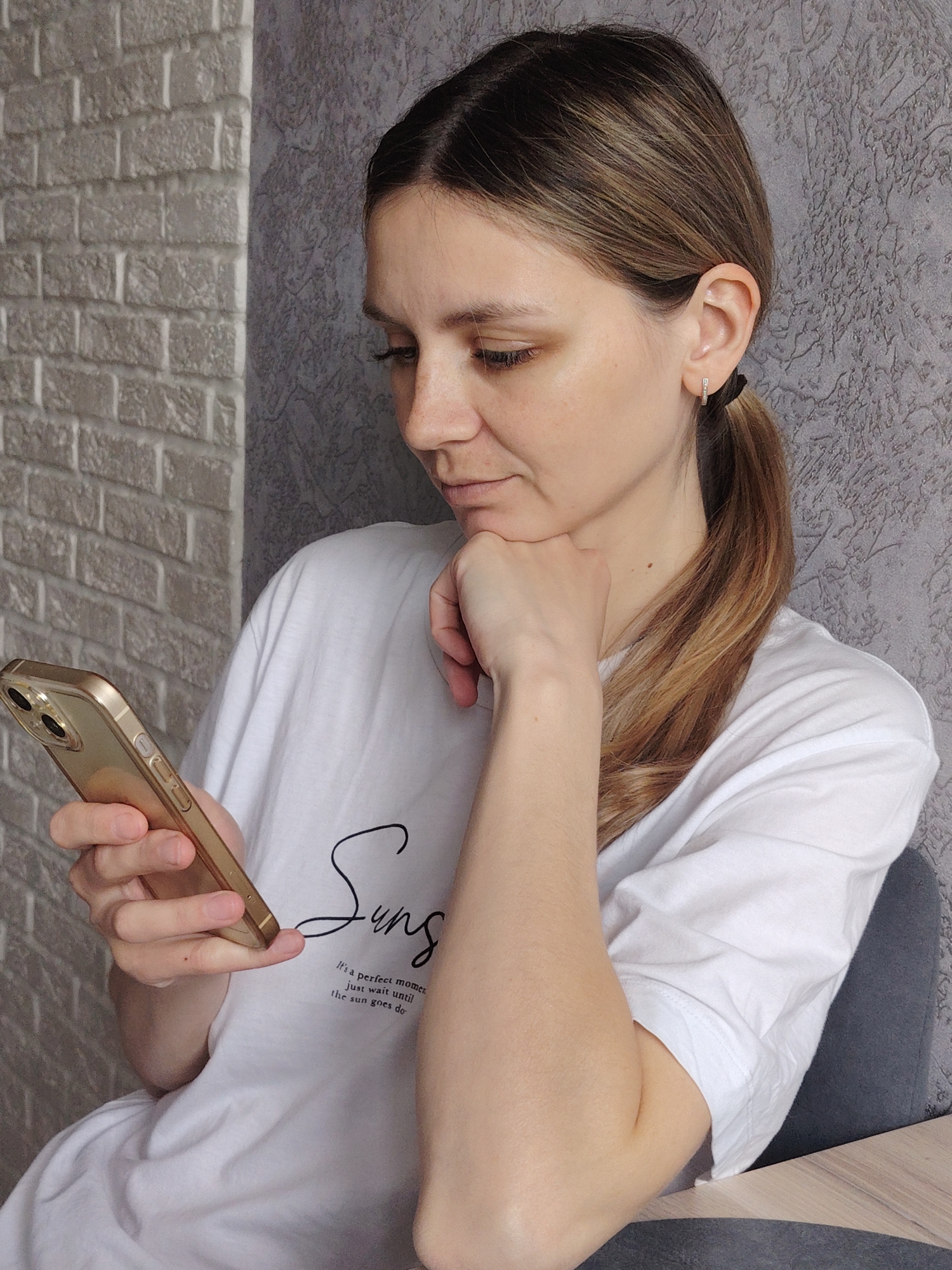 A woman is seated indoors, likely in a residence in Chaikovsky, Russia, based on the prompt. She holds a gold-colored mobile phone in her left hand, focusing her gaze on the screen. Her head rests slightly on her left fist. She wears a white t-shirt with black lettering and has light brown hair pulled back in a ponytail. She is wearing small hoop earrings. The background consists of a light gray brick wall on the left and a darker gray textured wall on the right.