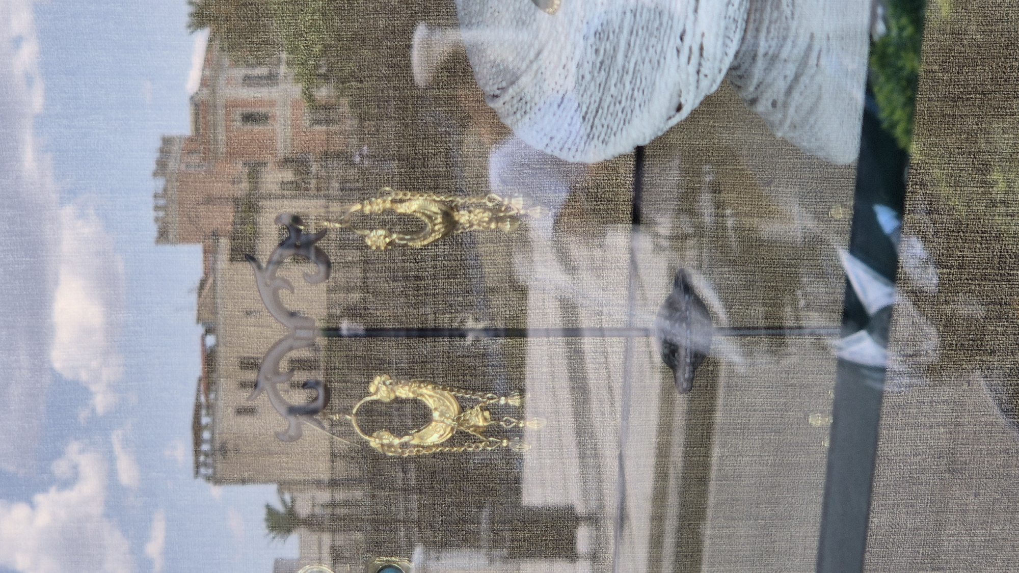 The image displays a pair of elaborate gold earrings hanging from a display stand within a storefront. The earrings are ornate, featuring a crescent-shaped top with dangling chains and small bead-like embellishments. Behind the earrings, a textured curtain or fabric is visible, partially obscuring a reflected scene. This reflection shows buildings and a cloudy sky, suggesting the storefront is situated in an urban environment. The architecture of the reflected buildings appears to be old, with towers and distinct window styles. The presence of greenery further indicates an outdoor setting. This scene is captured from outside a shop window, as evidenced by the slight glare and reflections. The overall impression is that of a jewelry shop window display, likely in a historical district of a city, possibly Taranto, Italy, given the architectural style which is reminiscent of some older Italian cities. There are no people visible in the scene, and no discernible actions are taking place.