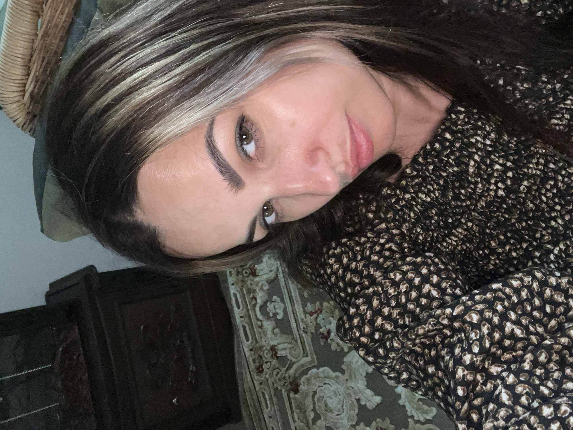 A close-up, low-angle, indoor shot shows a woman with brown and blonde streaked hair and brown eyes looking toward the camera. She is wearing a dark brown shirt with a leopard print of lighter brown and white spots. The background shows a dark wooden piece of furniture on the left, a patterned rug beneath it, and a wicker basket on the right.