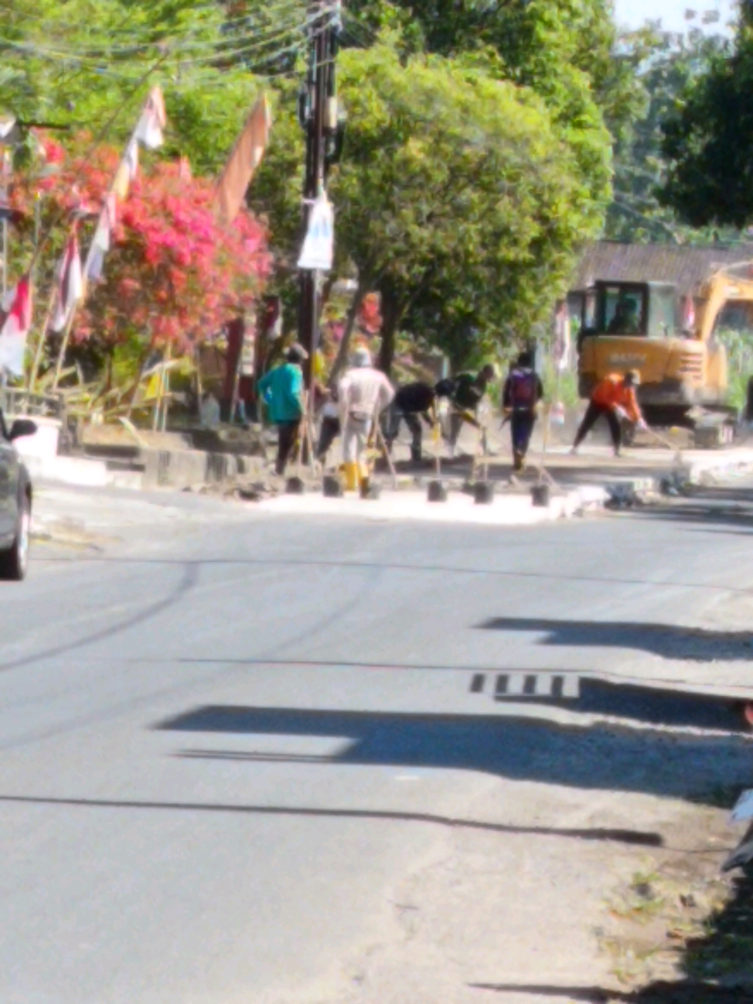 The image depicts a street scene in Sukorini, Indonesia, focusing on a road construction project. A yellow excavator is visible in the background. Several individuals are actively engaged in road work, some appearing to be shoveling or moving materials. One person is wearing a bright turquoise shirt, another an orange shirt, and others are dressed in more muted tones. A dark-colored car is partially visible on the left side of the frame. The environment includes trees and foliage, suggesting a semi-urban or rural setting. Flags and banners are also present, indicating a possible local event or celebration. The road surface appears to be undergoing repair or construction, with piles of white material scattered on the ground.