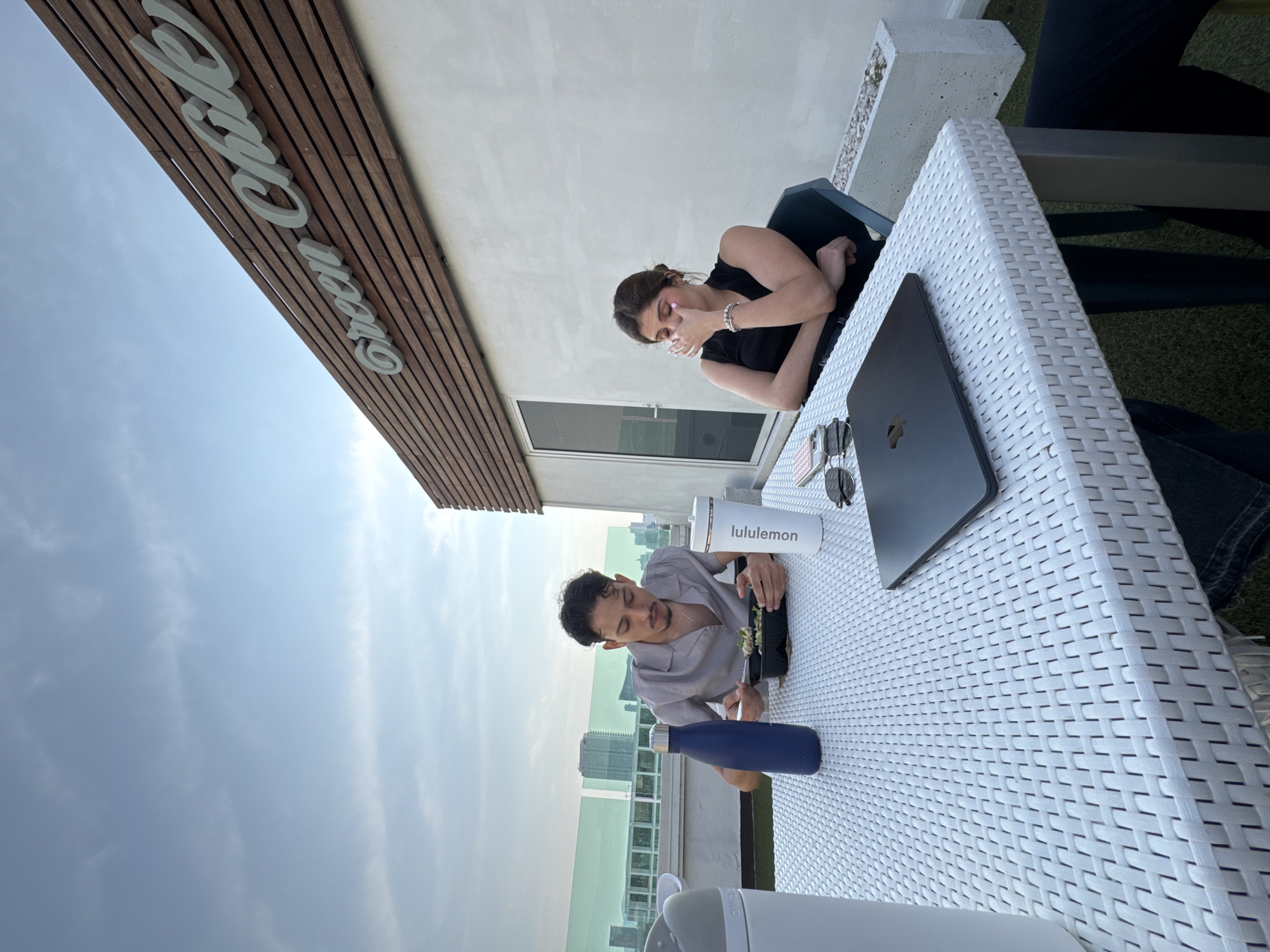 The image depicts two individuals seated at a white, lattice-patterned table at what appears to be an outdoor café. The man, located at the bottom of the frame, is wearing a light grey button-down shirt and is eating with a fork and container. A dark blue water bottle and a white "lululemon" bottle are on the table. The woman, located toward the top of the frame, is wearing a black sleeveless top and is touching her face with her right hand. A closed, black laptop, and a pair of sunglasses are on the table next to her. The scene is set on a high vantage point, suggesting a rooftop. A sign reads "Green Cafe" and the sky behind reveals urban buildings. The location is likely in Heroica Puebla de Zaragoza, Mexico, as the "Green Cafe" logo is visible in the photo. The background consists of the city skyline.