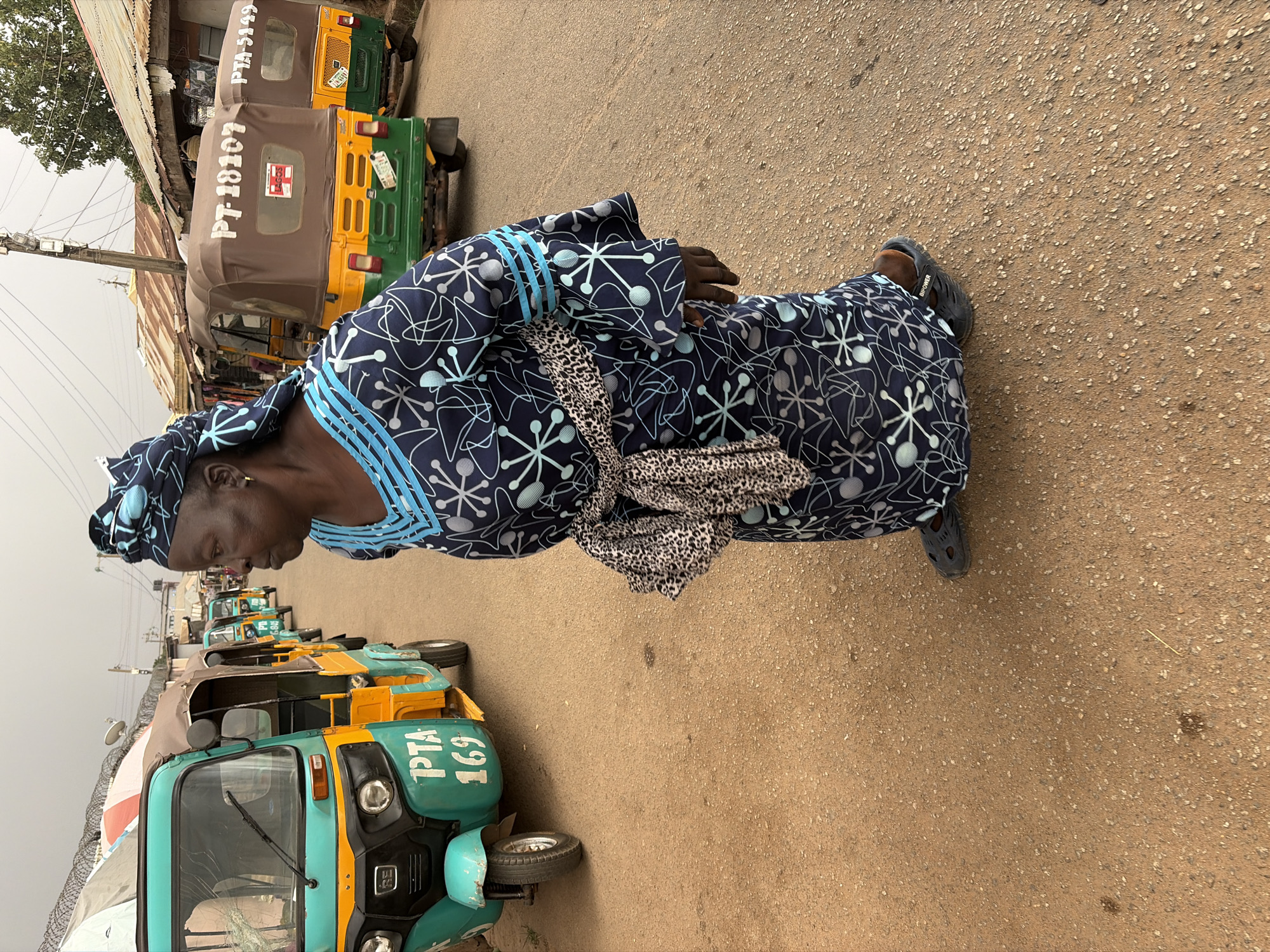 A woman is depicted standing or walking on a light brown, gritty street, likely in Jos, Nigeria. She is dressed in a vibrant navy blue attire featuring light blue and white abstract geometric patterns, complete with a matching headwrap. A leopard print sash or fabric is tied around her waist. She wears dark-colored slip-on shoes, one of which has "UNIPOWER" visible on its side. Her gaze is directed downwards, and her expression is neutral.
The background features a row of parked auto-rickshaws, locally known as "keke Napep," which serve as public transportation. These vehicles display varying color schemes, including yellow and green, and teal and yellow, with some having brown canopies. Visible text on the keke Napeps includes "PT-18107" and a "LAGOS" sticker on a brown-canopied vehicle, "PTA-5949" on another in the distance, and "PTA 169" on a teal and yellow one closer to the foreground.
Beyond the vehicles, buildings with corrugated iron roofs are partially visible, suggesting an urban or semi-urban setting. A utility pole with wires stands to the left. The sky is overcast, indicating daytime. The scene captures a moment of daily life on a bustling street.