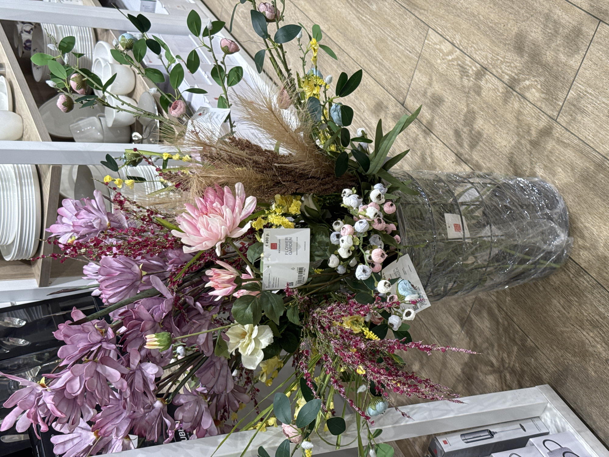 The image presents a vibrant artificial flower arrangement, likely displayed for sale in a retail store. The arrangement is composed of a diverse selection of faux flowers and foliage, featuring prominent large pink and purple chrysanthemum-like blossoms, clusters of small white, pink, and light blue flowers, and delicate sprigs of tiny yellow and dark red/maroon blooms. Lush green leaves and stems provide a verdant base, complemented by feathery brown elements resembling pampas grass.
The flowers are held in a dark, possibly metallic or plastic, vase with a geometric pattern, which is wrapped in clear protective plastic. The arrangement sits on a surface, with a light brown wooden floor visible to the right.
In the background on the left, white shelves are stocked with various white ceramic dishes, including plates, cups, and bowls, suggesting a home goods section.
Several white price tags are attached to the arrangement. One clearly displays "1 490 p.", indicating a price in Russian Rubles, along with the brand "VILLEROY & BOCH" and the product name "FLOWER GARDEN." A barcode is also visible on this tag. Another tag partially seen also shows "FLOWER GARDEN." The scene is brightly lit, typical of an indoor retail environment.