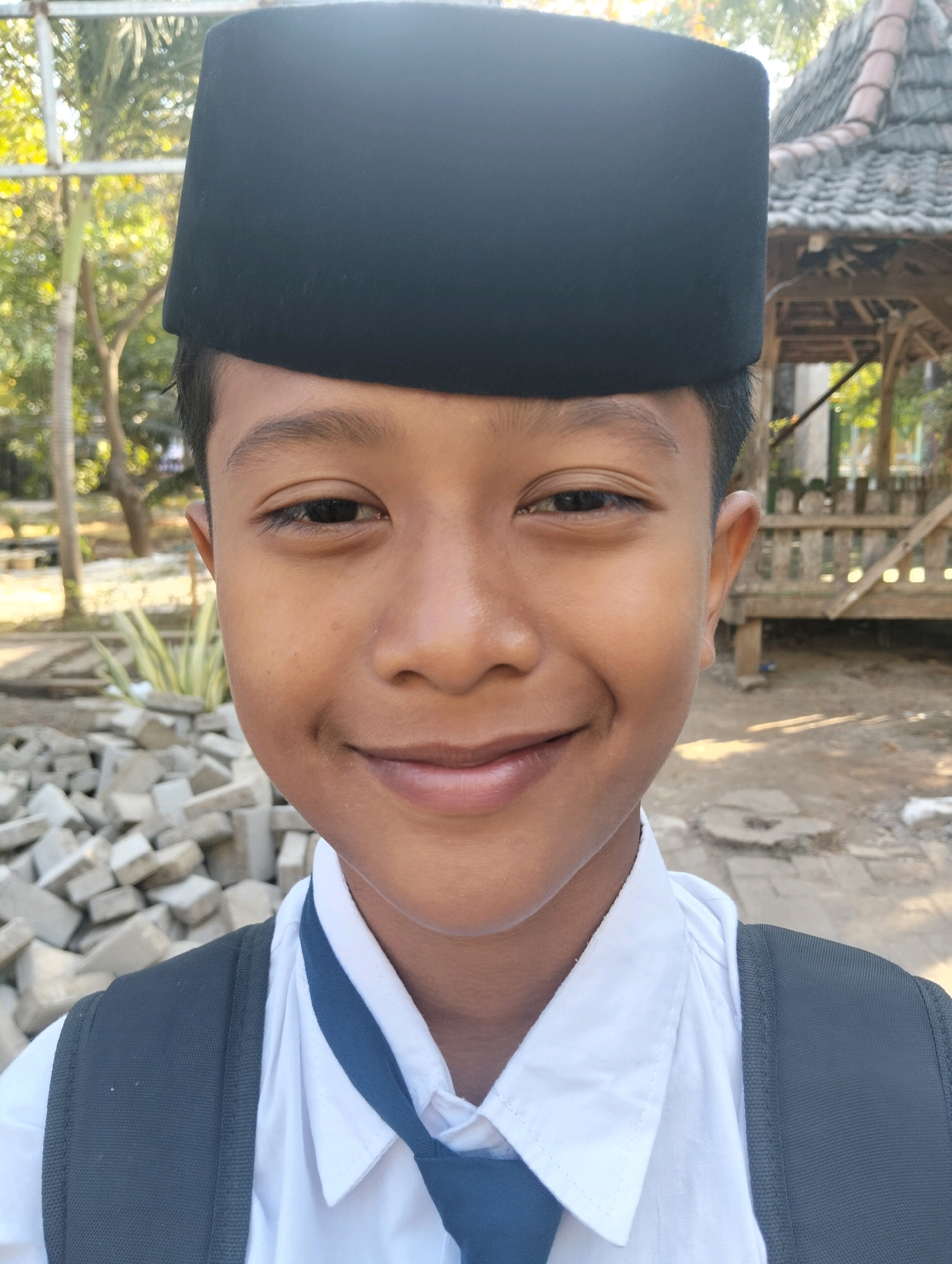 A young boy is pictured from the chest up, facing the camera directly with a slight smile. He is wearing a black peci, a white collared shirt with a blue tie, and a grey backpack. The background is slightly out of focus, but appears to be outdoors in a sunny environment with trees and a rustic wooden structure. The ground is visible in the foreground, covered with scattered bricks. The image is likely taken in Paciran, Indonesia, based on the architecture and vegetation commonly found in the region.