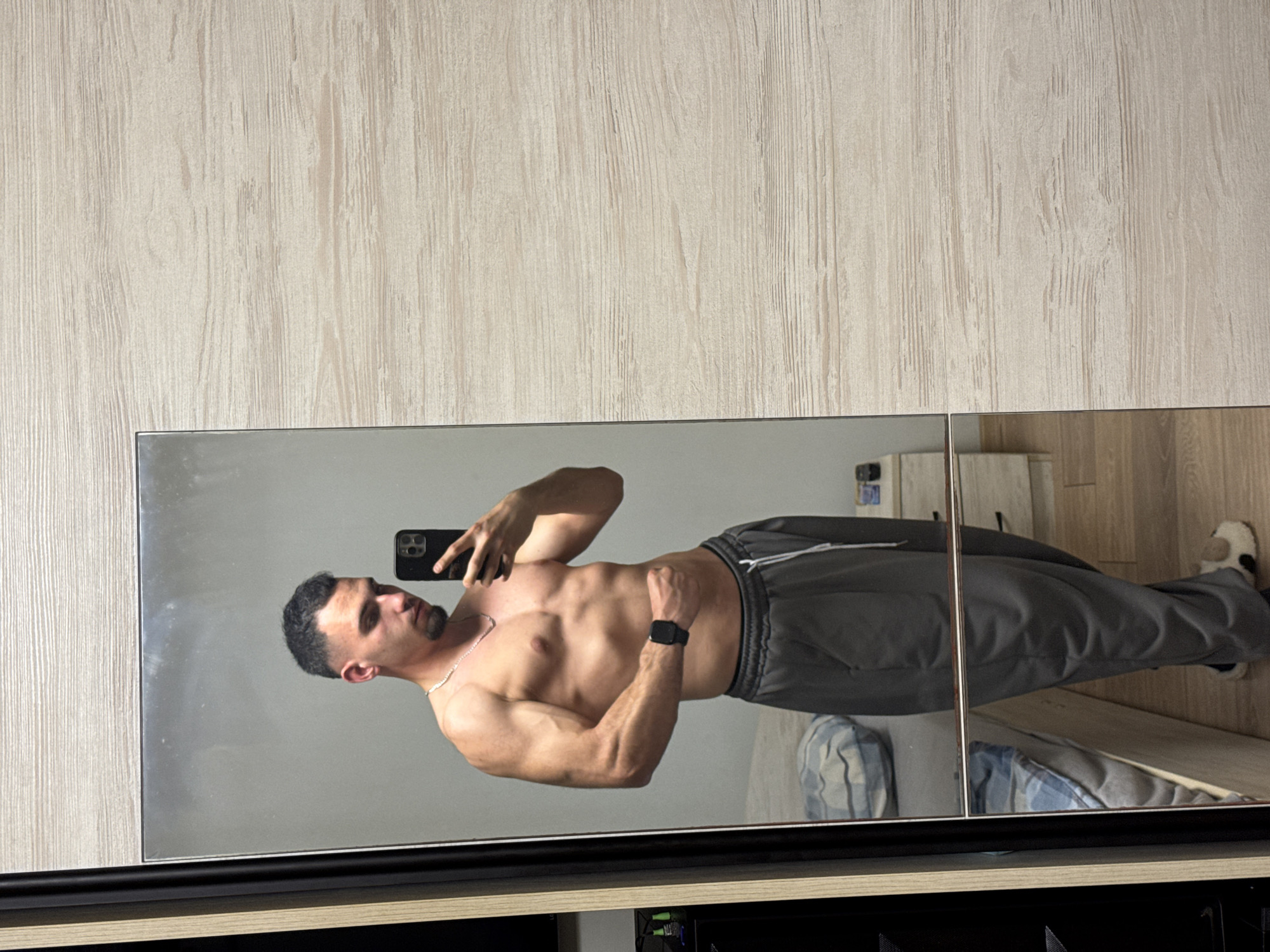A shirtless man is taking a selfie in front of a mirror. The man is wearing grey sweatpants and a smartwatch. The background shows a wooden wall and a cabinet. The location is not specified.