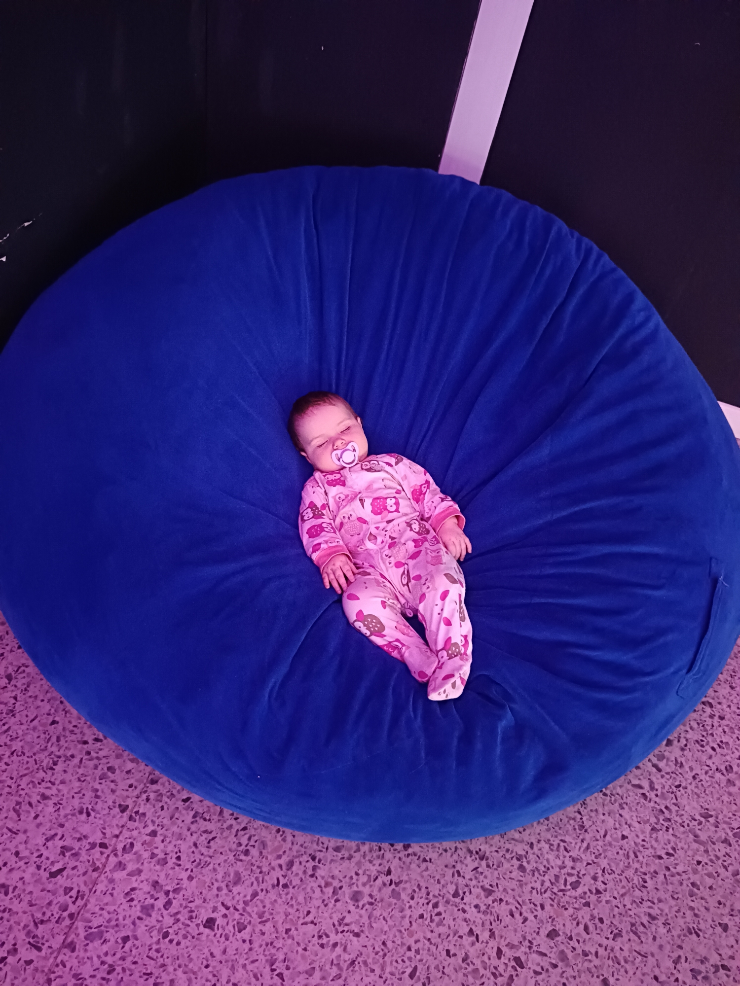 An overhead shot captures a baby peacefully sleeping on a large, round, royal blue bean bag chair. The infant is clad in a pink footed sleeper patterned with owls and has a white and pink pacifier in its mouth, with eyes closed in repose. The bean bag appears soft and plush, showing natural folds in its fabric.
The bean bag is situated on a floor characterized by a distinctive speckled or terrazzo pattern in shades of pinkish-purple. In the background, a dark wall, possibly black or dark grey, is visible, intersected by a narrow, light-colored vertical strip, which could be a beam or decorative element, on the right side. The ambient lighting subtly casts a purple hue across the floor and parts of the bean bag.
The scene is indoors, conveying a sense of calm and comfort. No visible text is present in the image. The location context for this image is Chaikovsky, Russia.