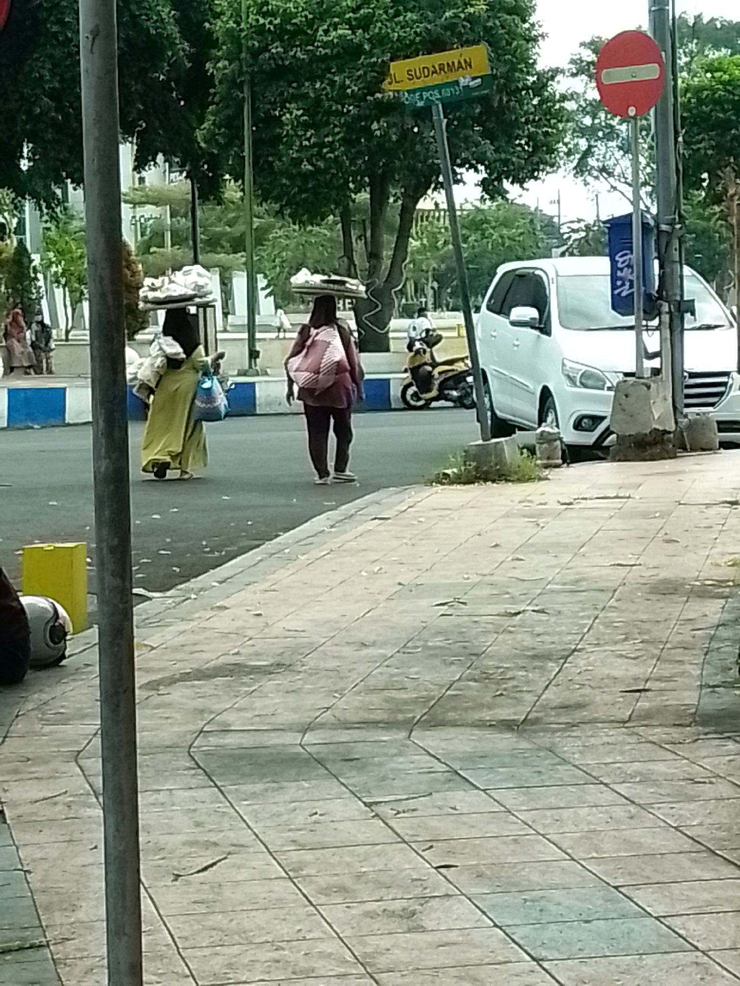 The image shows a street scene in Jember, Indonesia. A yellow street sign reads "JL. SUDARMAN" and "ODE POS. 68131". A red traffic sign with a white horizontal bar is visible. Two women are walking on the street. One woman is wearing a yellow dress and carrying items on her head. The other woman is wearing a dark top and pants, carrying a pink backpack. A white car is parked on the right side of the road. A motorcycle is parked on the left side of the road. A yellow rectangular object is on the sidewalk. The sidewalk is paved with gray tiles. Trees and buildings are visible in the background.