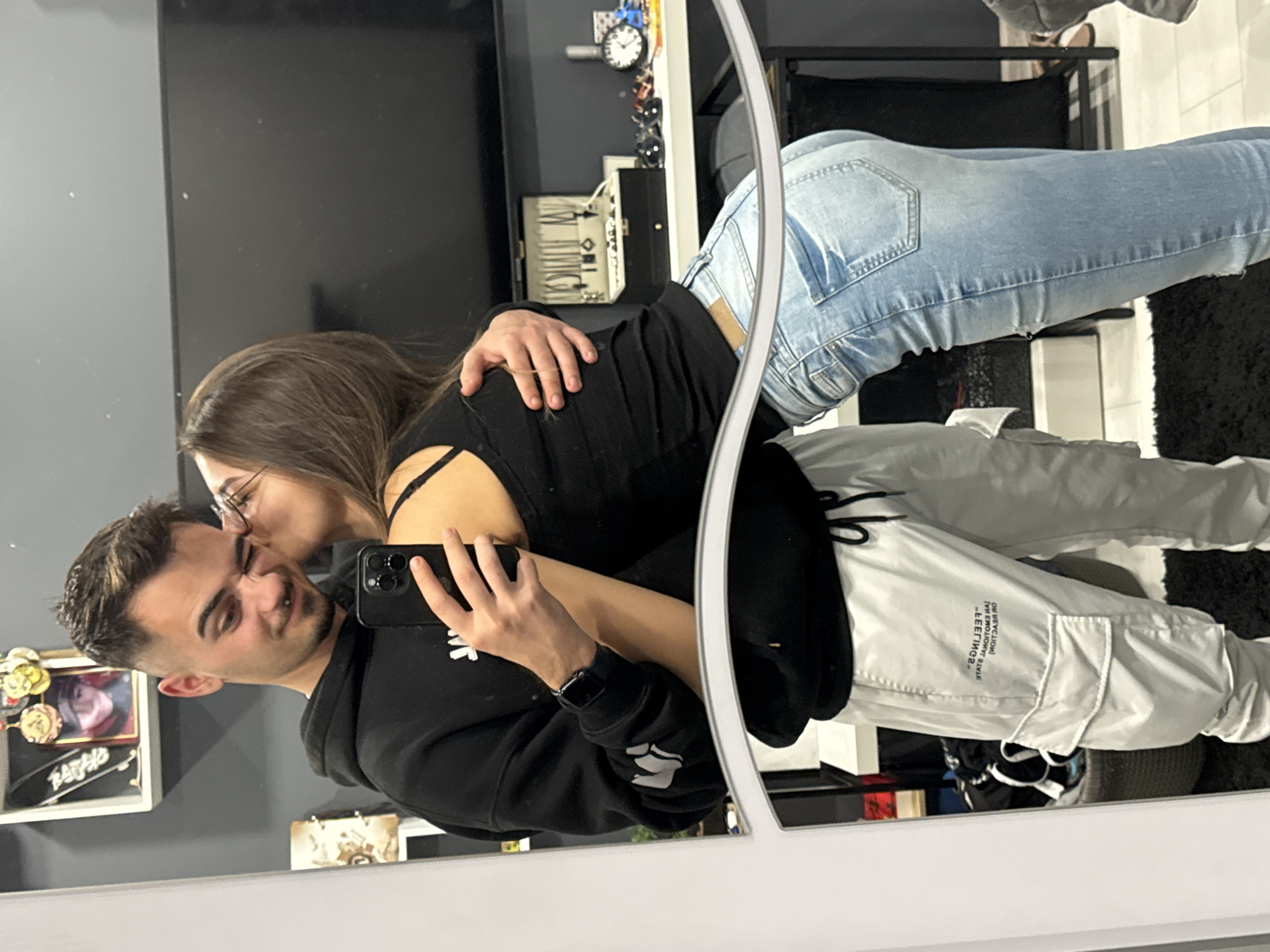 A couple is taking a selfie in a mirror. The man is holding the phone, and the woman is kissing him on the cheek. The man is wearing a black hoodie and white pants, and the woman is wearing a black top and blue jeans. The background includes a black wall and a shelf with various items, including a clock and jewelry. The location is likely indoors, but not definitively within the city of Sofia, Bulgaria, based on visual evidence alone.