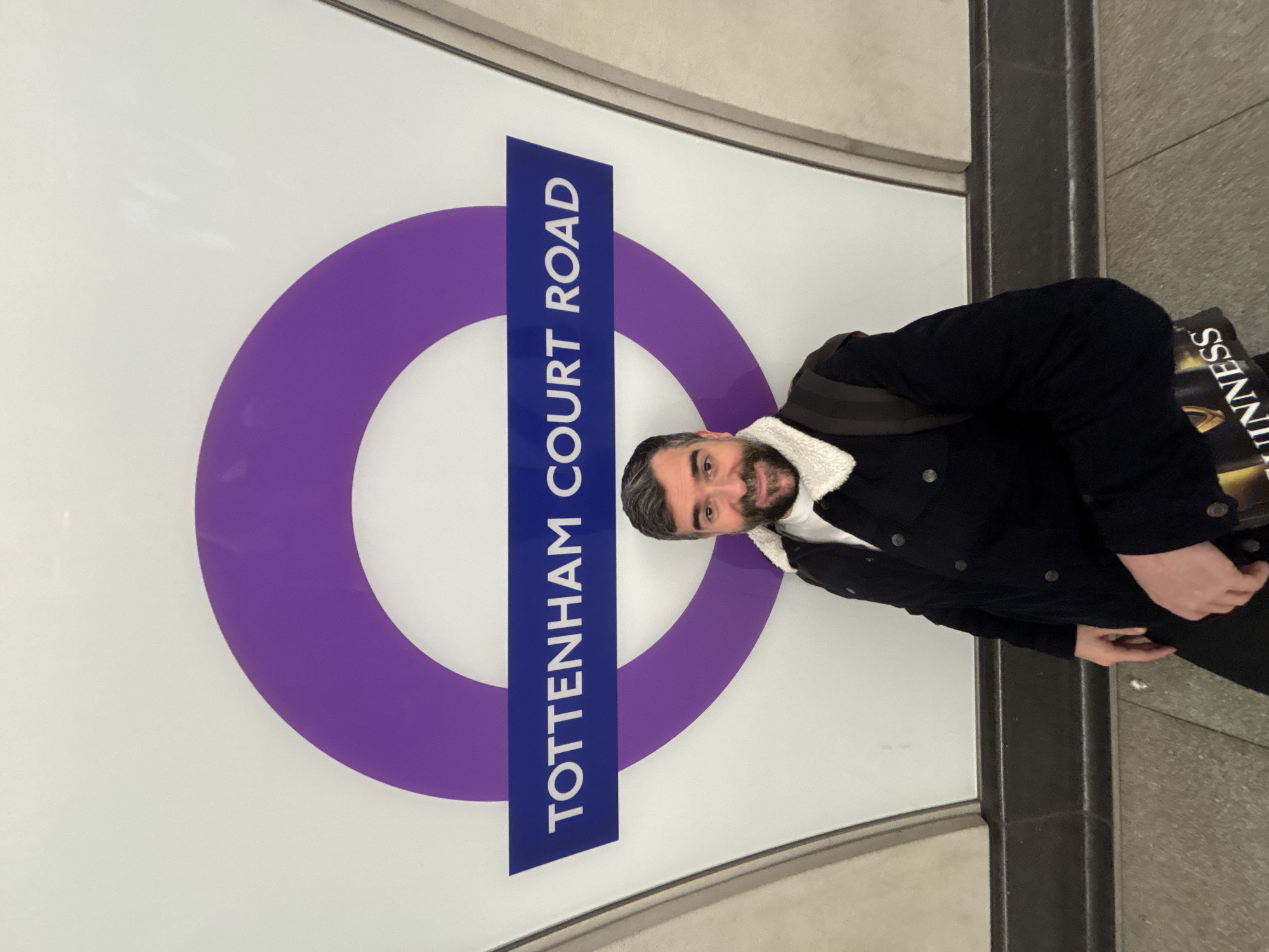 A man stands in front of the Tottenham Court Road station sign on the London Underground. He is wearing a dark blue jacket with a white, sherpa-like collar and a dark backpack. He has dark hair and a beard. The sign is a purple circle with a blue bar across the center, reading "Tottenham Court Road." The background appears to be the station platform, with gray tiled walls and floors. The man is slightly rotated towards the right, and the sign is oriented vertically.