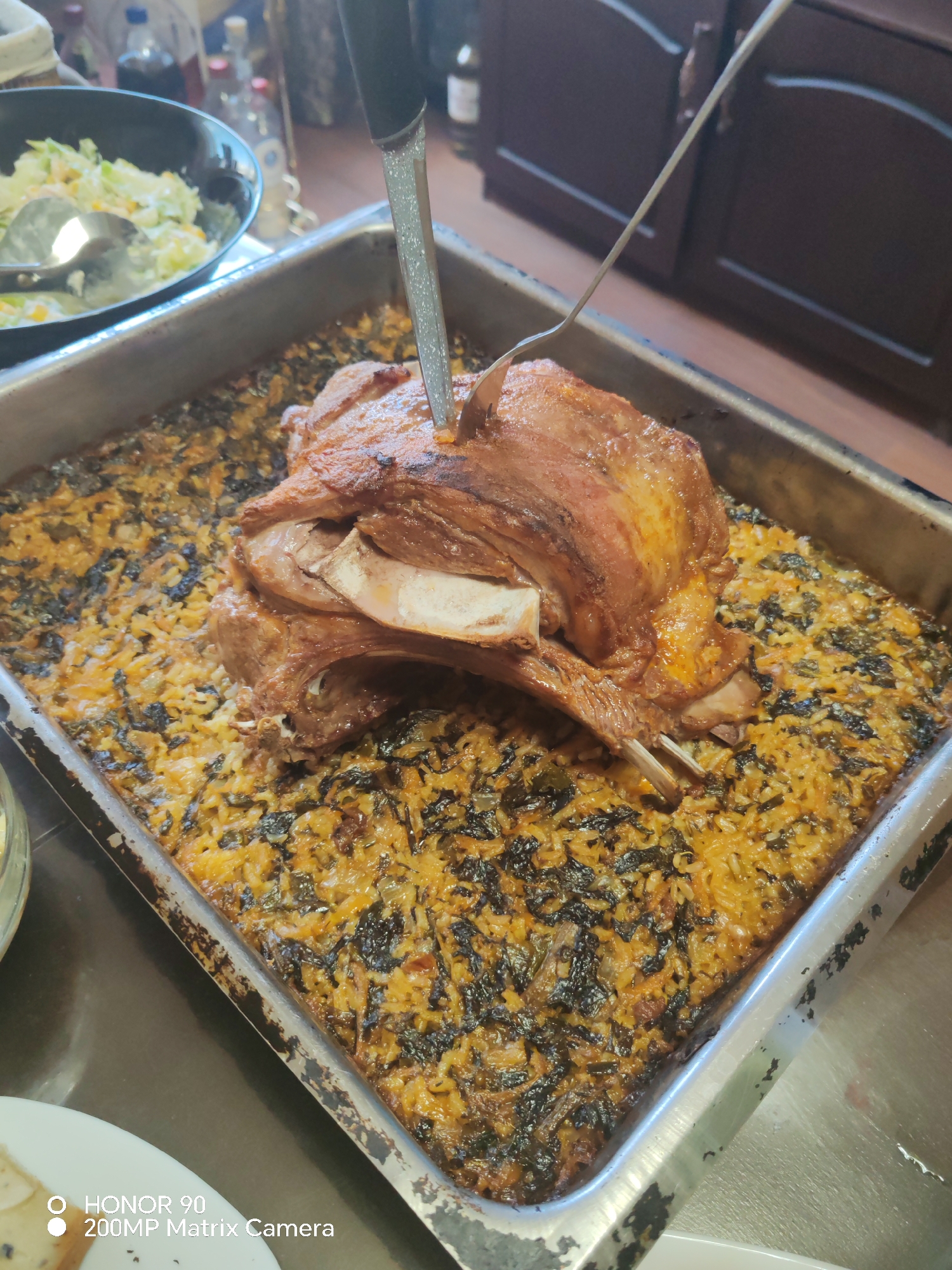 A close-up shot captures a roasted meat dish in a rectangular metal tray. The meat, possibly lamb, is positioned centrally on a bed of rice and spinach. A carving knife is inserted into the meat, and a two-pronged fork rests on the right side of the meat. In the upper left corner, a bowl containing a salad is partially visible, along with bottles of liquids. The tray sits on a stainless steel surface. The environment suggests a kitchen or dining area. The location in the city of Sofia, Bulgaria, is not deducible from the visual information.
