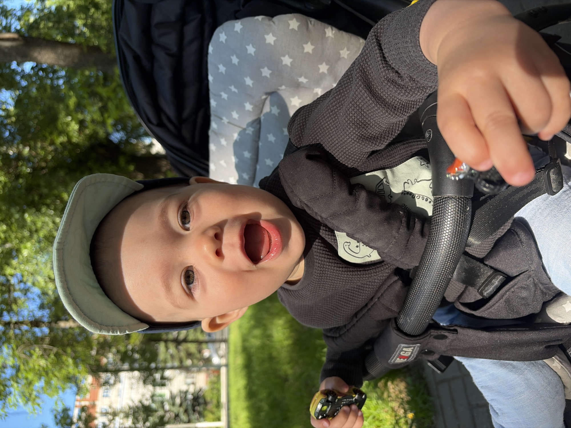 The image shows a close-up of a baby sitting in a stroller outdoors on a sunny day. The baby is wearing a light blue cap and a black long-sleeved shirt with a graphic design. The baby has an open mouth and appears to be smiling or laughing. The baby is holding a small toy in their right hand, and another toy is visible in their left hand near the stroller handle. The stroller has a black frame and a grey cushion with white stars. In the background, there is a green grassy area, trees, and a building. The scene is likely in Chaikovsky, Russia, given the mention of the city. The overall environment suggests a park or residential area.