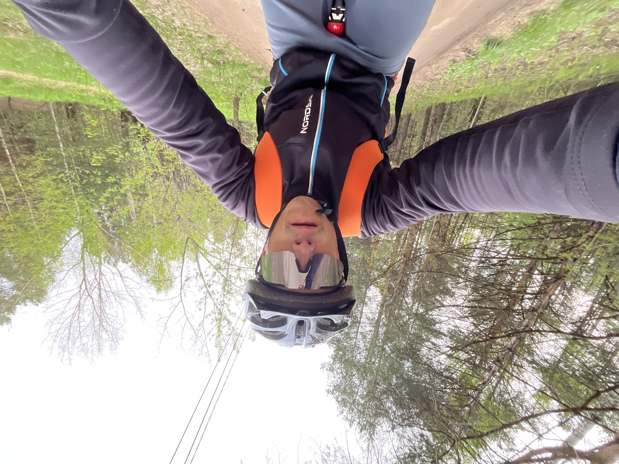 A cyclist is taking a selfie while riding on a dirt trail. The camera angle is looking up at the cyclist, who is wearing a helmet and sunglasses, and a cycling jacket with orange shoulder panels and black sleeves. The jacket has the word "NORDDEK" on the chest. The cyclist is riding on a dirt path with trees on either side. The location is likely Chaikovsky, Russia.