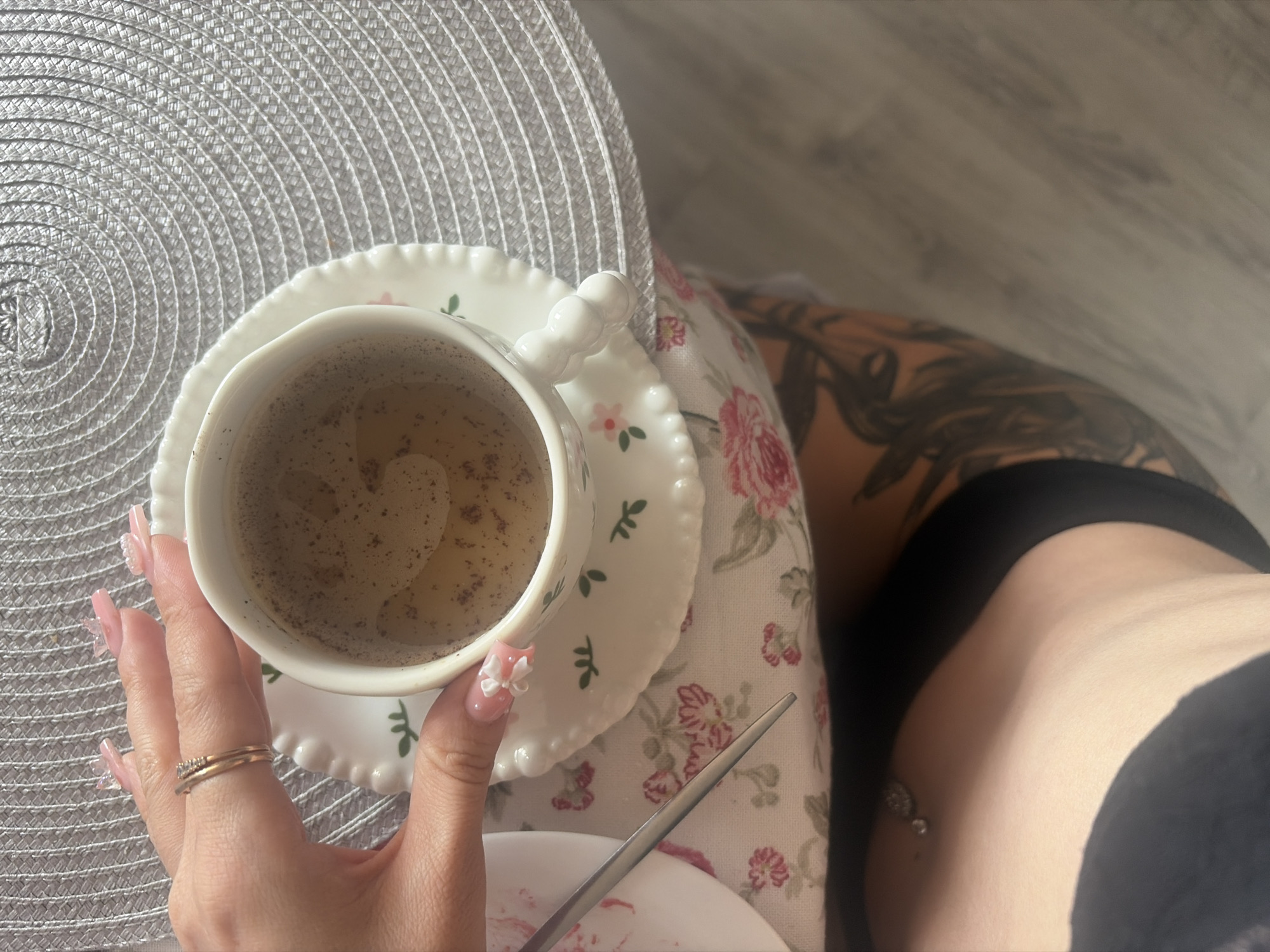 An overhead shot captures a detailed view of a table setting and parts of a person. A fair-skinned hand, adorned with long, pink nails featuring delicate 3D white bow and flower embellishments and small pearls, holds a white ceramic cup with a distinctive rounded handle. The cup contains a light brown liquid, possibly coffee or tea, with small dark particles floating on the surface. The cup rests on a matching white saucer with a scalloped edge, decorated with small green leaf motifs and tiny pink flowers. On the ring finger of the hand, two gold rings are worn: a plain band and a thinner band with small embedded diamonds.
Beneath the cup and saucer is a round, light gray woven placemat featuring concentric circular patterns. This placemat is positioned on a white tablecloth adorned with a floral pattern of large pink roses and green foliage. To the left, a portion of another white plate with pink residue and a silver utensil, possibly a spoon or knife, is visible.
Below the table level, the upper abdomen of the person is visible, featuring a navel piercing with an ornate, possibly jeweled, silver top ball. Further down, a thigh with a large, dark, intricate tattoo, likely botanical in nature, is partially visible, suggesting the person is wearing dark clothing, possibly shorts. The background shows a light-colored floor with subtle wood grain patterns. The image provides no specific visual cues to determine its location within the city of Saint Petersburg, Russia, or any other geographical area.