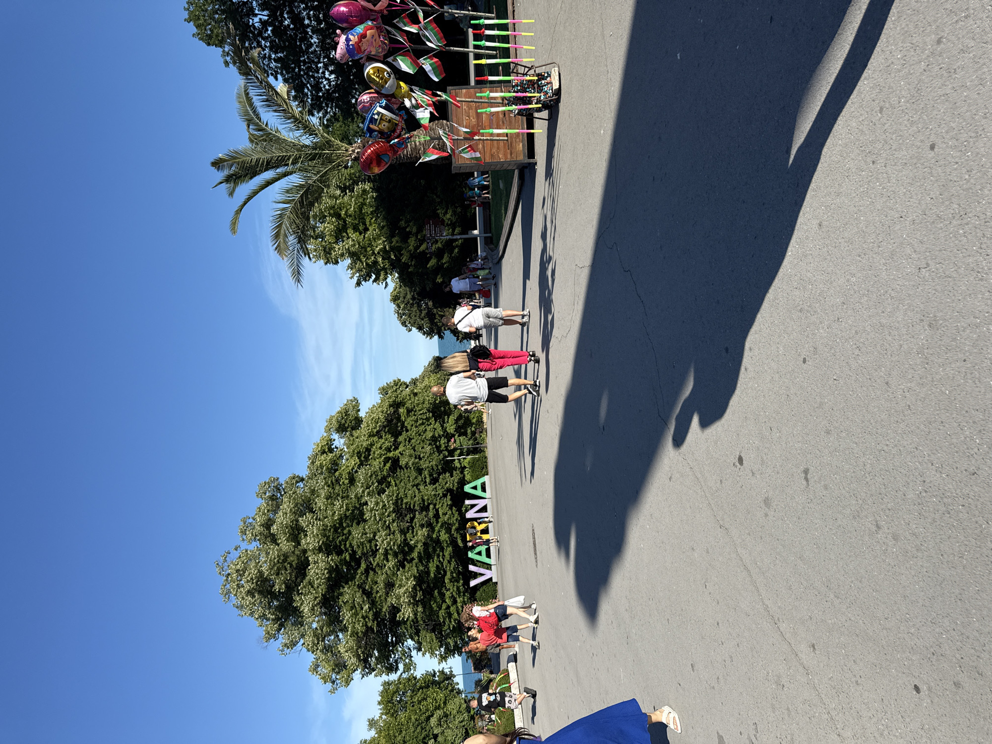 This media file shows an outdoor scene on a sunny day. Several people are walking along a paved path, some individually and some in small groups. In the background, there are trees and a clear blue sky. To the left, a stall is set up with a variety of balloons and flags on sticks, suggesting a festive atmosphere or a vendor selling souvenirs. A shadow of what appears to be a person or people is cast on the ground, indicating a bright, sunny time of day, likely midday. In the mid-ground, large, colorful, three-dimensional letters spell out "VARNA". This text is a strong indicator of the location. The overall mood seems relaxed and leisurely, with people enjoying an outdoor activity in what appears to be a park or promenade.