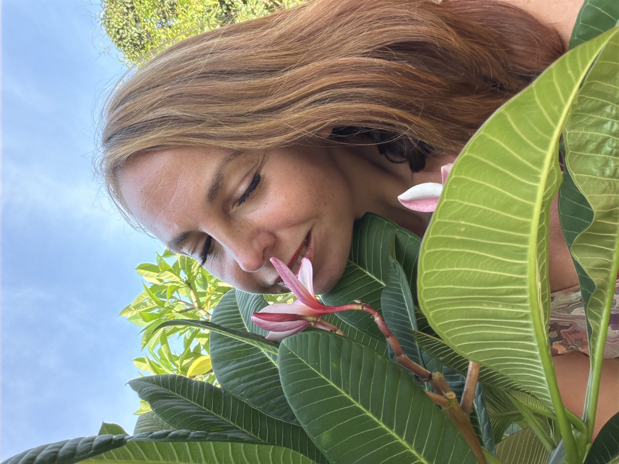 The image features a woman with long, light brown hair, looking down and slightly to the left, with her eyes closed as she smells a pink and white flower. She is surrounded by lush green leaves of a plant, likely plumeria, given the flower's appearance and the tropical foliage. The scene is set outdoors, with a bright blue sky visible in the background, suggesting a sunny day. The angle of the shot is from below, looking up at the woman and the plant. Her expression conveys a sense of peace and enjoyment of the moment. The lighting suggests it is daytime, possibly late morning or early afternoon. The woman's skin has a warm tone, and she is wearing a garment with a colorful, abstract pattern, partially visible. The focus is on her face, the flower, and the surrounding leaves, creating an intimate and serene atmosphere. The location context of Baan Mae Nam, Thailand, implies a warm, tropical environment. There is no visible text in the image.