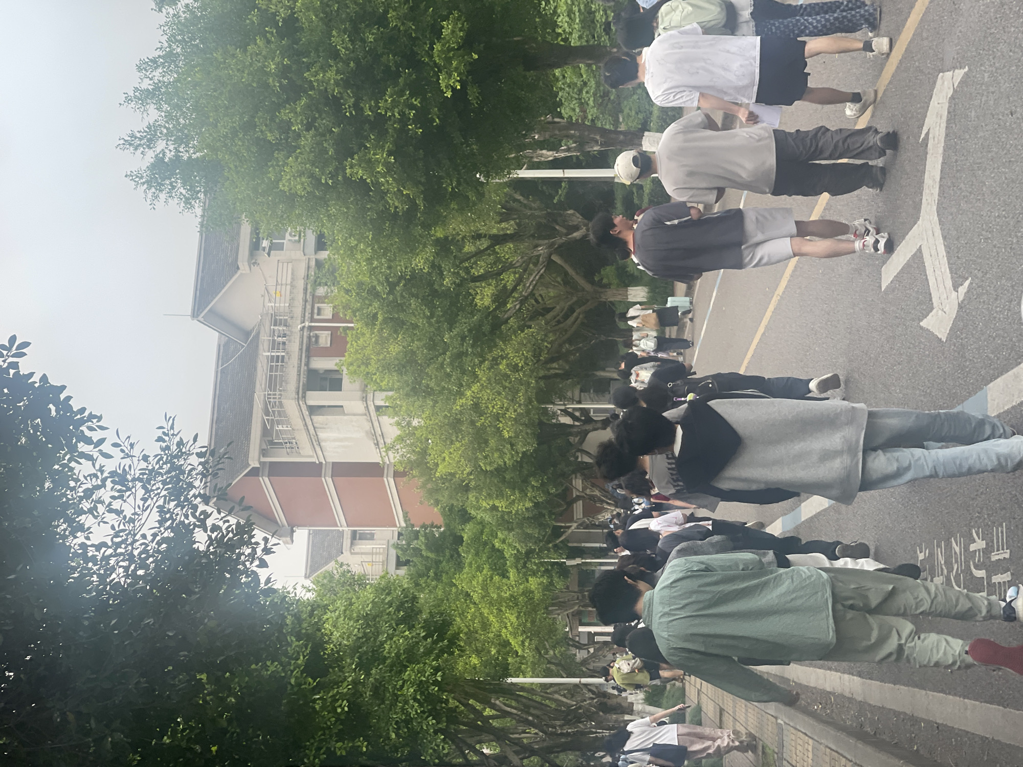 The image shows a group of people standing in a line on an asphalt road. The location is Chongqing High-tech Zone, China, indicated by the Chinese characters painted on the road. A multi-story building with a combination of red brick and white facade is visible in the background, partially obscured by green trees lining the road. The individuals in the line vary in attire. The scene appears to be an outdoor gathering.