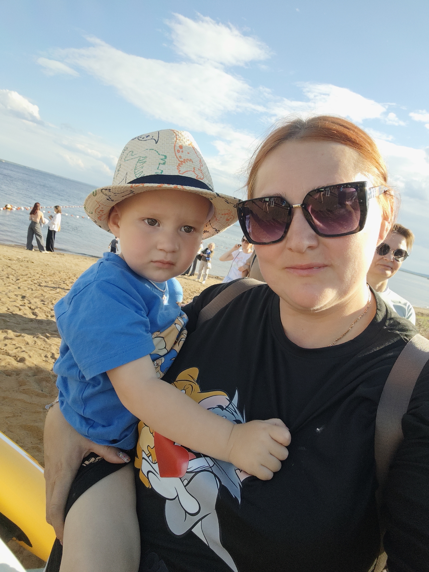The image is a selfie taken at a beach. The main subjects are a woman and a young child she is holding. The woman is wearing a black t-shirt with a cartoon graphic and large sunglasses. She has reddish-blonde hair. The child is wearing a blue t-shirt and a patterned fedora-style hat. The child's expression is serious.
In the background, there are other people walking on the sandy beach and near the water. The sea is visible, and there are buoys in the water. The sky is blue with white clouds, suggesting it is a sunny day. The overall setting is a recreational beach, likely during daylight hours.
The image was taken in Chaikovsky, Russia, as indicated by the location context. There is no visible text in the image. The scene conveys a casual, perhaps family outing to the beach.