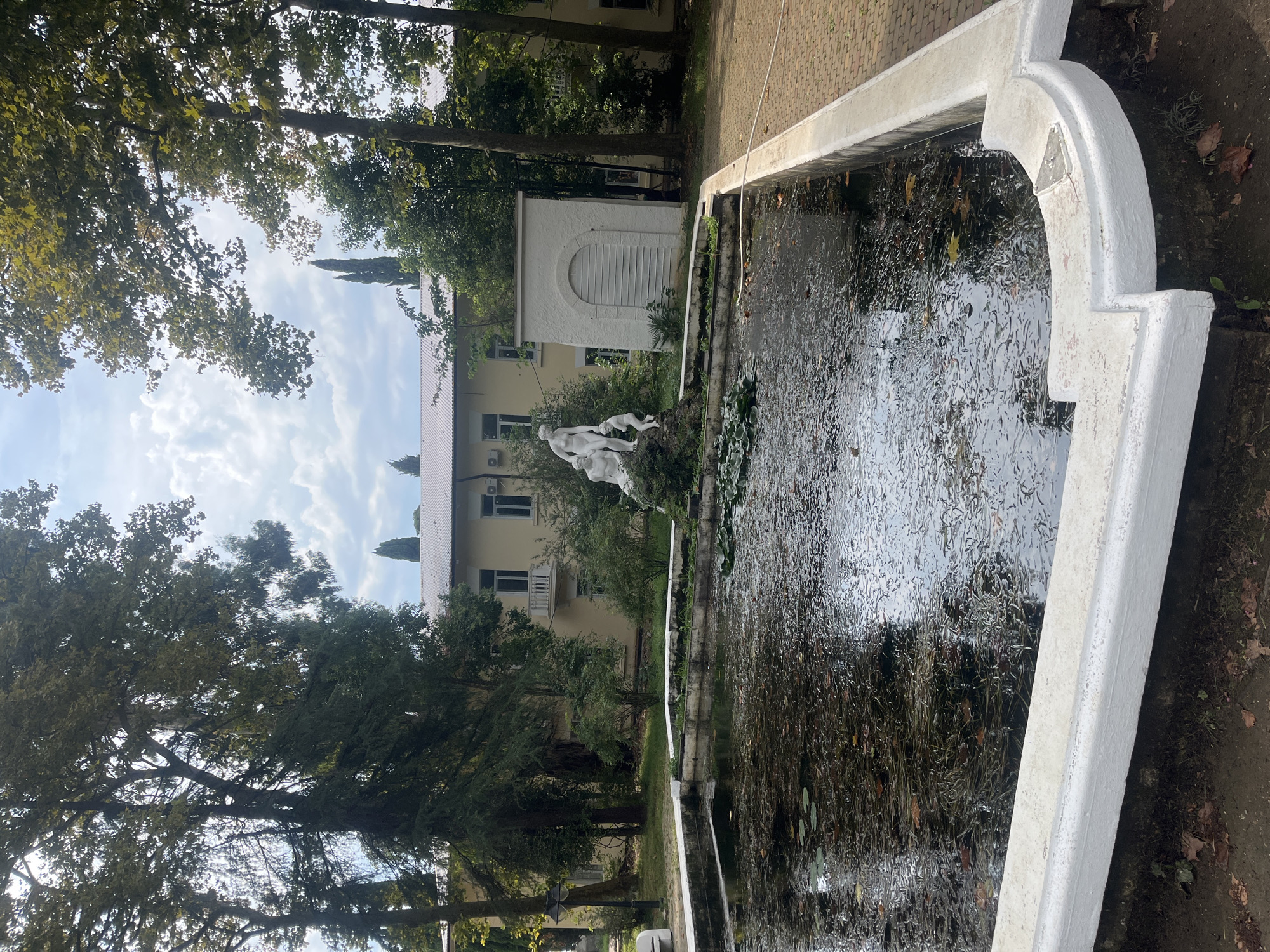 The image depicts a rectangular pond with a white concrete rim in a park-like setting. The pond water is dark and contains numerous fallen leaves and aquatic plants. In the center of the pond, a sculptural group of three white figures, two adult and one child, is situated on a rocky mound covered in greenery. Behind the pond, a light yellow building with a white roof is visible, surrounded by mature trees. To the right of the building, a small white structure with a rounded, louvered door is present. The surrounding area includes a paved walkway made of reddish-brown bricks and patches of grass. The sky above is partly cloudy. The scene appears to be in the Arboretum in Sokhumi, Abkhazia.
