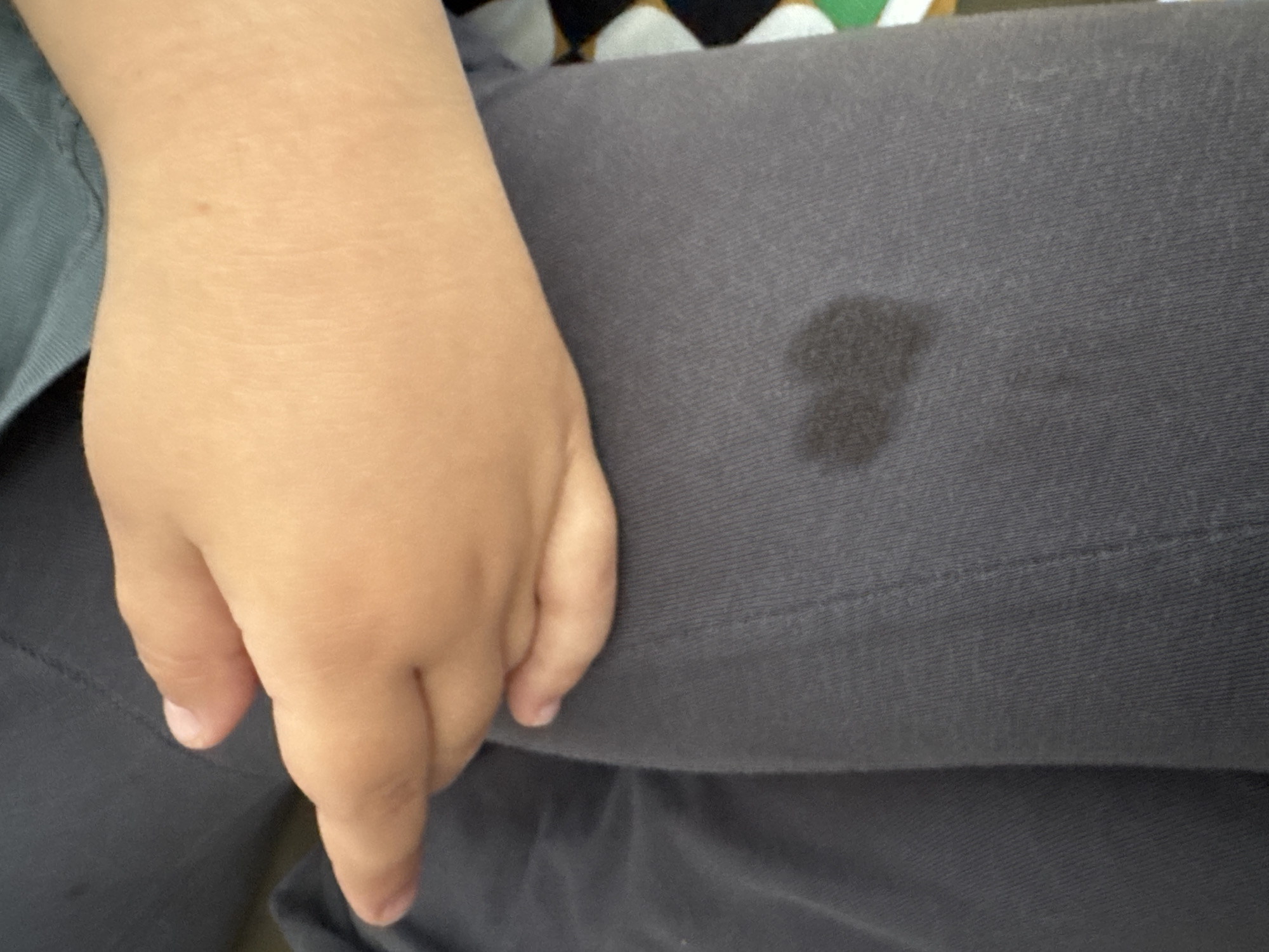 A child's hand rests on a person's lap, which is covered in gray fabric. There is a dark, wet spot on the fabric near the hand. The location is unspecified, it does not specify if it is in Sofia, Bulgaria.