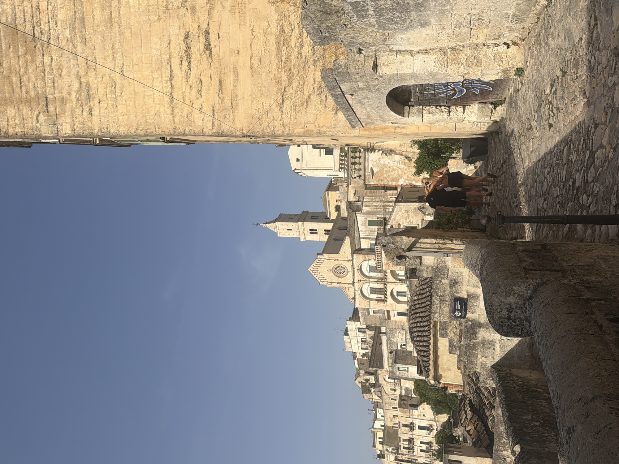 The image depicts a view of the Sassi di Matera, a cave dwelling district in the city of Matera, Italy. In the foreground, two individuals, a male and a female, walk away from the camera down a cobblestone path. The male is wearing a black t-shirt and shorts, while the female is wearing a white top and dark shorts. They are walking past a textured stone wall on the right side of the image, which features an arched doorway with a weathered wooden door decorated with blue graffiti. Further back, a dense cluster of ancient stone buildings with terracotta tiled roofs fills the landscape, ascending a hillside under a clear blue sky. A prominent bell tower, characteristic of Matera's architecture, rises above the buildings in the distance. The sunlight casts shadows, highlighting the textures of the stone structures and the uneven terrain.