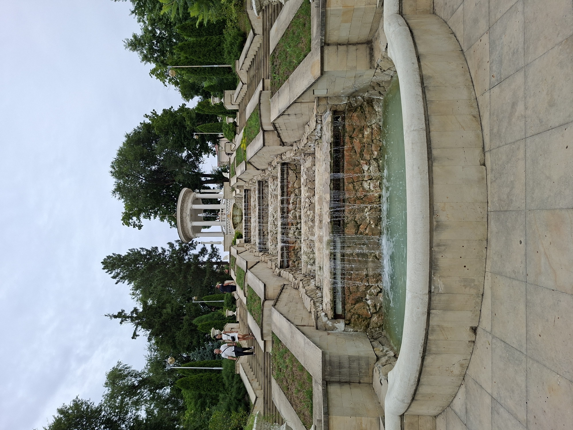 The image shows a cascading fountain with multiple tiers of stone and water, set against a backdrop of lush green trees and an overcast sky. Several people are visible on the wide, stone staircase that flanks the fountain, walking up and down. The fountain is constructed with rough-hewn stone and concrete, featuring water flowing over several levels into a large, curved pool at the bottom. The architecture of the fountain is grand and formal, with symmetrical steps and decorative urns placed on ledges. Above the fountain, at the top of the steps, a white, open-air pavilion or gazebo with columns can be seen. The environment is a park or public garden, likely in a city setting, given the man-made structures. The specific location is the Cascade fountain in Chișinău, Moldova, a well-known landmark.