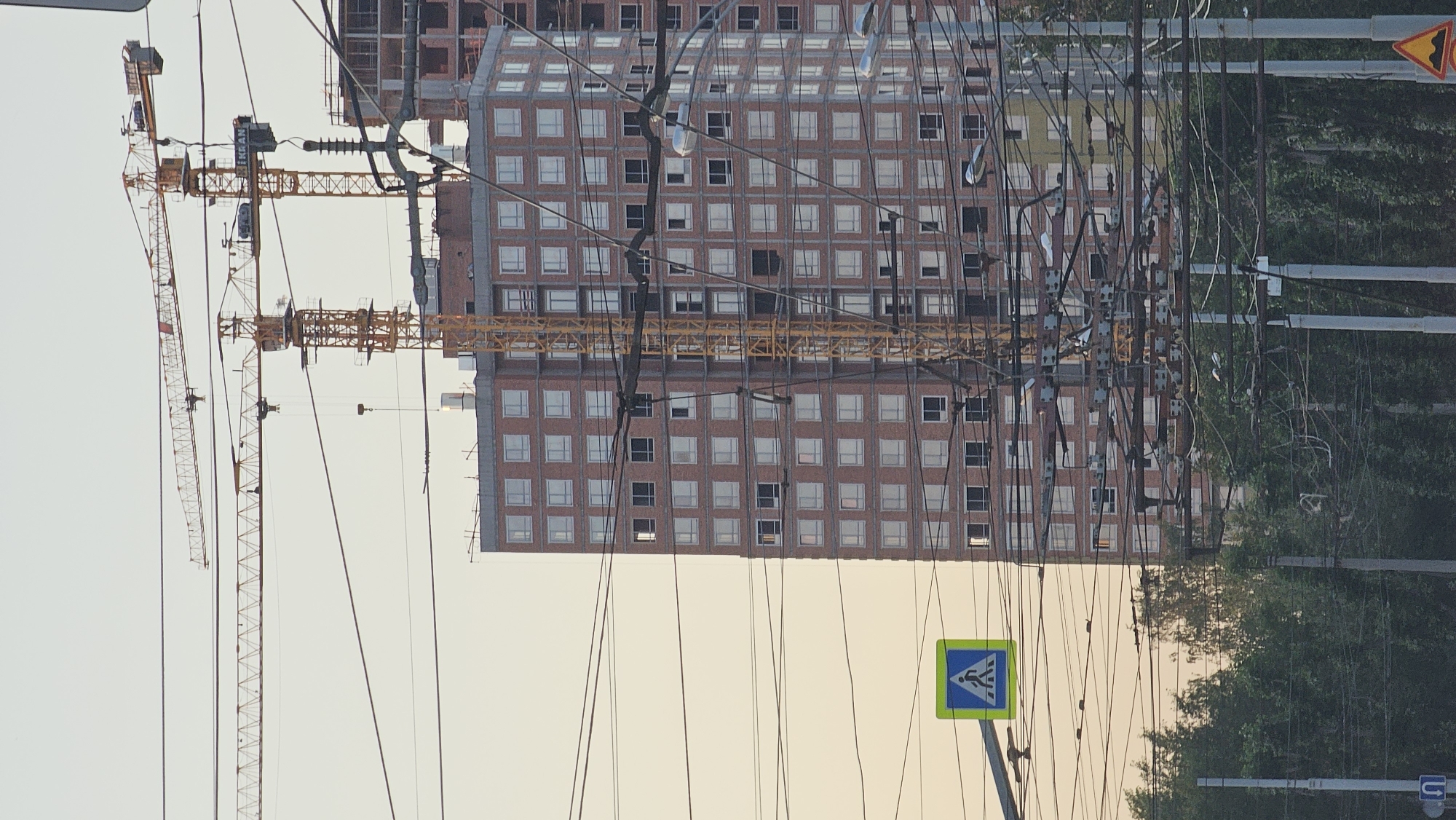 The image captures a construction scene in Yekaterinburg, Russia, featuring unfinished multi-story buildings and two yellow construction cranes. The building in the foreground is clad in red brick with many windows, some of which are dark, indicating they are not yet fitted with glass. A pedestrian crossing sign is visible in the lower left quadrant of the image, a common sight in urban environments. A dense network of overhead electrical wires crisscrosses the scene, obscuring parts of the buildings and cranes, suggesting the area is a well-trafficked urban thoroughfare. Trees line the lower edge of the image, forming a green contrast to the man-made structures. The sky is a pale, hazy orange, possibly indicating dawn or dusk. There are no people clearly visible in the image. The overall impression is one of ongoing urban development.