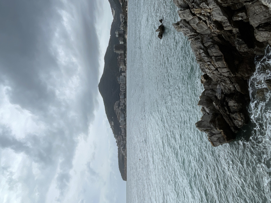 The image depicts a coastal scene in Budva, Montenegro, under a heavily overcast sky. In the foreground, dark grey, jagged rock formations extend into the water from the lower left and right, with white seafoam visible where waves impact them. The midground consists of a body of greenish-blue water with visible ripples and small waves. A small cluster of dark rocks is partially submerged in the mid-distance towards the center-right, and a tiny, dark, unidentified object floats on the water to the left. The background is dominated by a prominent, dark green, conical mountain, its slopes densely covered with buildings of the city of Budva. A slender communication tower is visible at the mountain's summit. The urban development stretches along the coastline, including multi-story structures and a visible beach section on the far right. The sky is entirely filled with thick, grey cloud formations. No individuals are visible.
