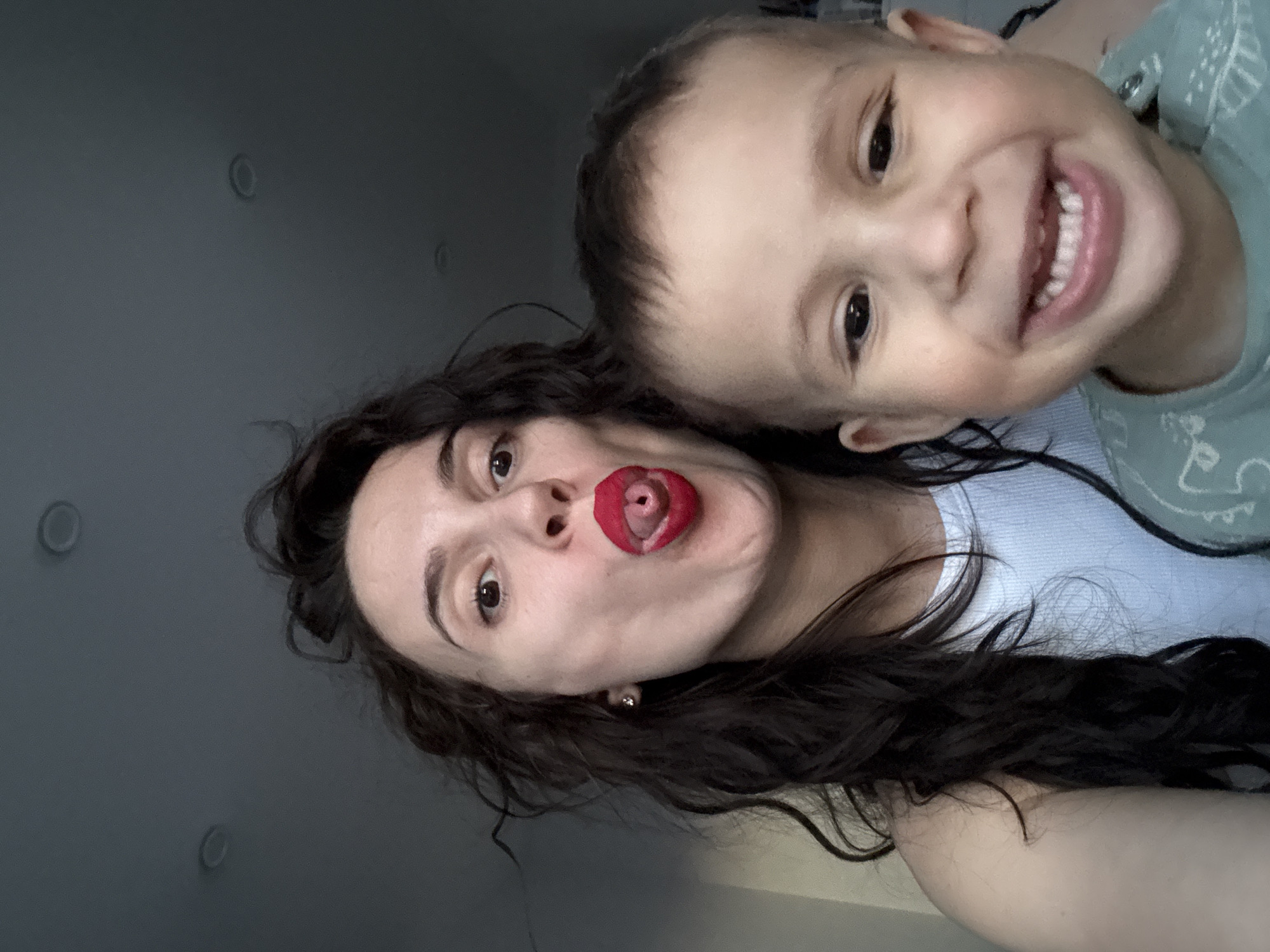 This vertical selfie captures a playful moment between a woman and a young child, likely indoors in Chaikovsky, Russia. The woman, positioned closer to the camera, has dark, wavy hair and is wearing a light-colored top. She is making a humorous face, sticking out her tongue, which is curled, and her lips are covered in a bold red lipstick. A small earring is visible in her right ear. Her dark eyes are looking directly at the camera.
Above and slightly behind her, a young child smiles broadly, revealing a full set of teeth. The child has fair skin and dark eyes, wearing a light-colored top with a subtle pattern. Both subjects appear to be in good spirits, conveying a sense of joy and affection.
The background is a plain, dark gray surface, possibly a wall or ceiling, with two circular indentations that could be recessed light fixtures. The lighting appears soft and even, suggesting an indoor setting without direct sunlight. There is no visible text in the image.