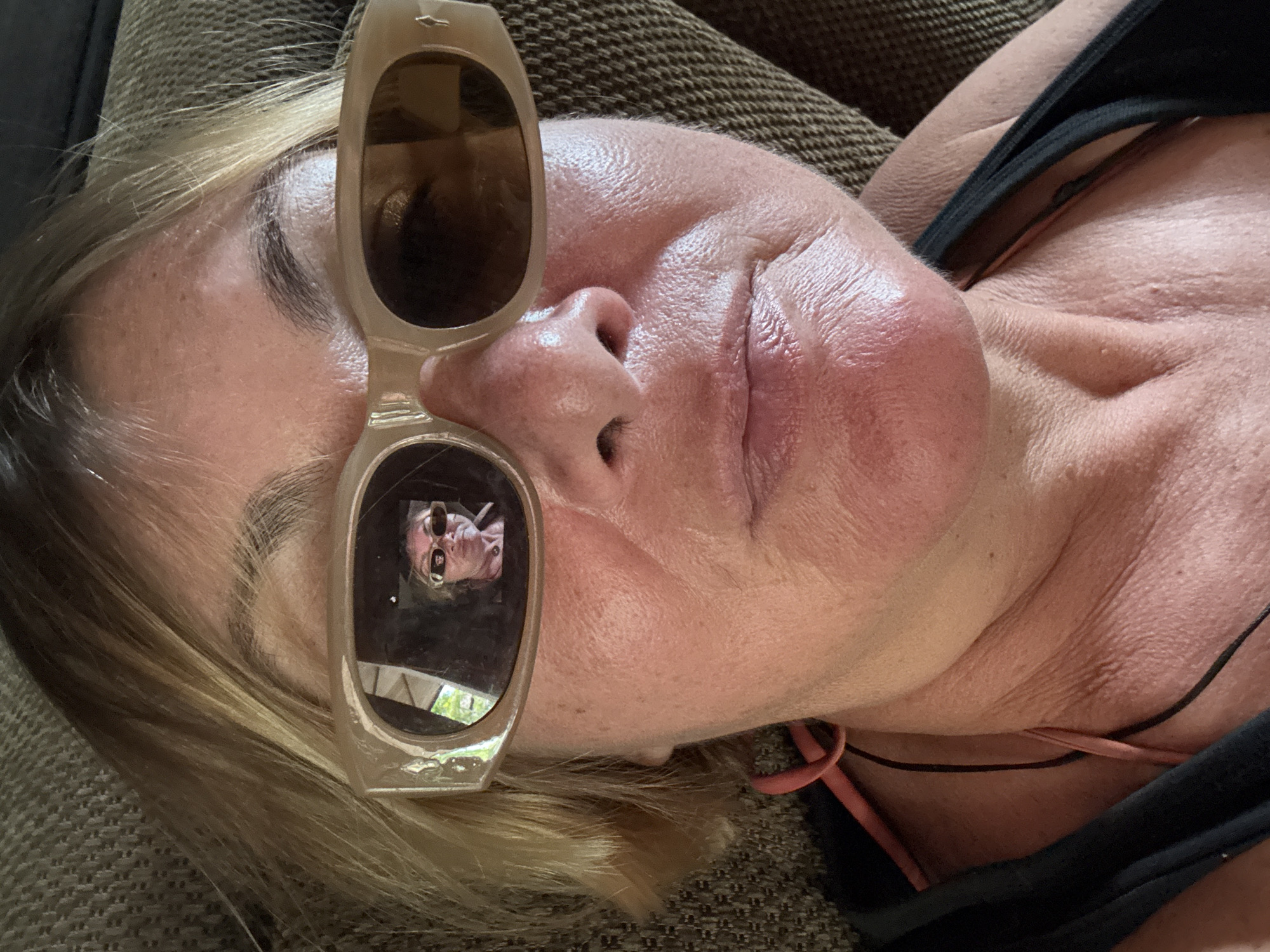 A close-up, selfie-style photo shows a woman from the shoulders up, wearing sunglasses that reflect another person. The woman has blonde hair and is wearing a black tank top with orange straps. The reflected person in the sunglasses is also wearing sunglasses and appears to be smoking. The background is a brown, textured surface, possibly a couch. The lighting suggests an outdoor or well-lit indoor environment.