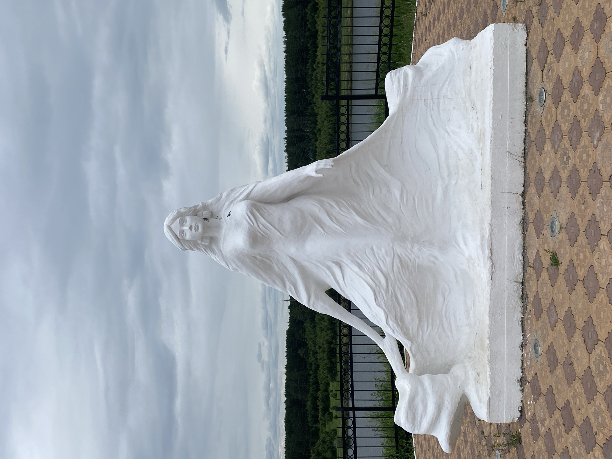 A white marble statue of a woman in a flowing gown stands on a patterned stone platform. The woman has long, wavy hair and her arms are outstretched, with her gown spread wide around her. In the background, there is a body of water, a dense forest, and a cloudy sky. The statue is located in Novomaltinsk, Russia.