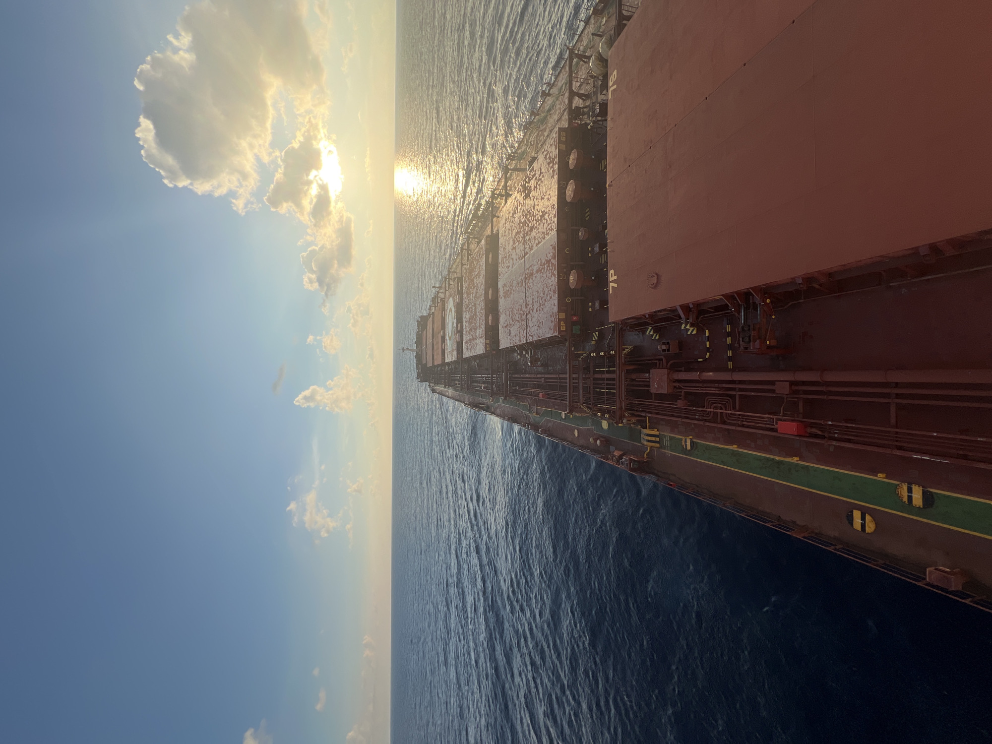 An aerial view looks down on the deck of a large cargo ship sailing on a calm sea under a bright sky. The sun is setting or rising on the right side of the image, casting a golden glow on the water and the ship's cargo hatches. The deck is largely covered by large, flat, red-brown hatches, some of which appear slightly wet. Along the left side of the deck, there is a walkway with green and yellow safety markings. The ocean stretches out to the horizon, with a gentle swell. The sky is a pale blue with scattered white clouds, some of which are illuminated by the sun. The ship is moving away from the viewer and towards the horizon. There are no people visible in the image.