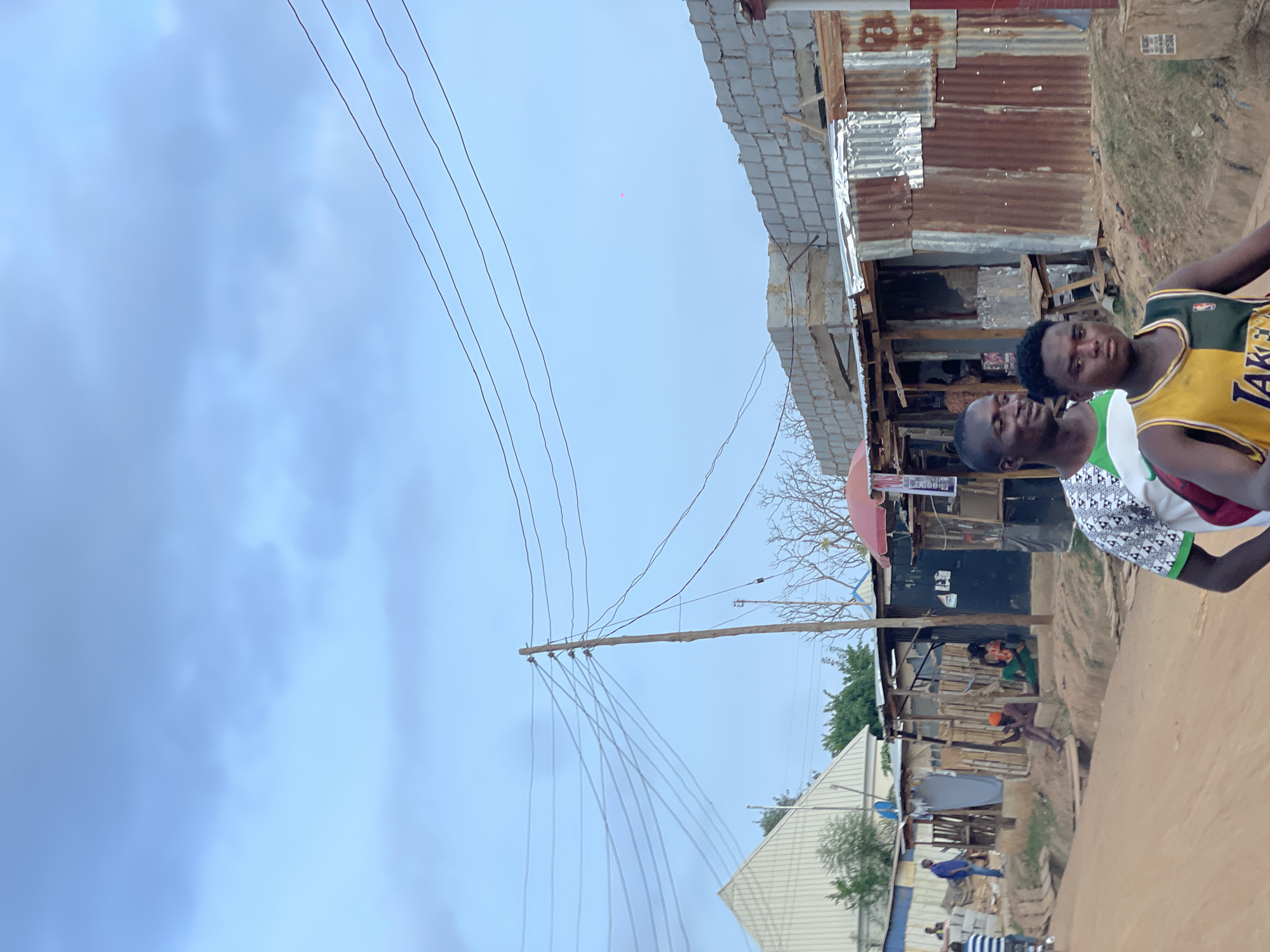 Two young men stand on a dirt road next to shacks in Karmajiji, Nigeria. Power lines run overhead. One man wears a yellow and green Lakers jersey. In the background, people work amid a construction site with corrugated metal shacks.