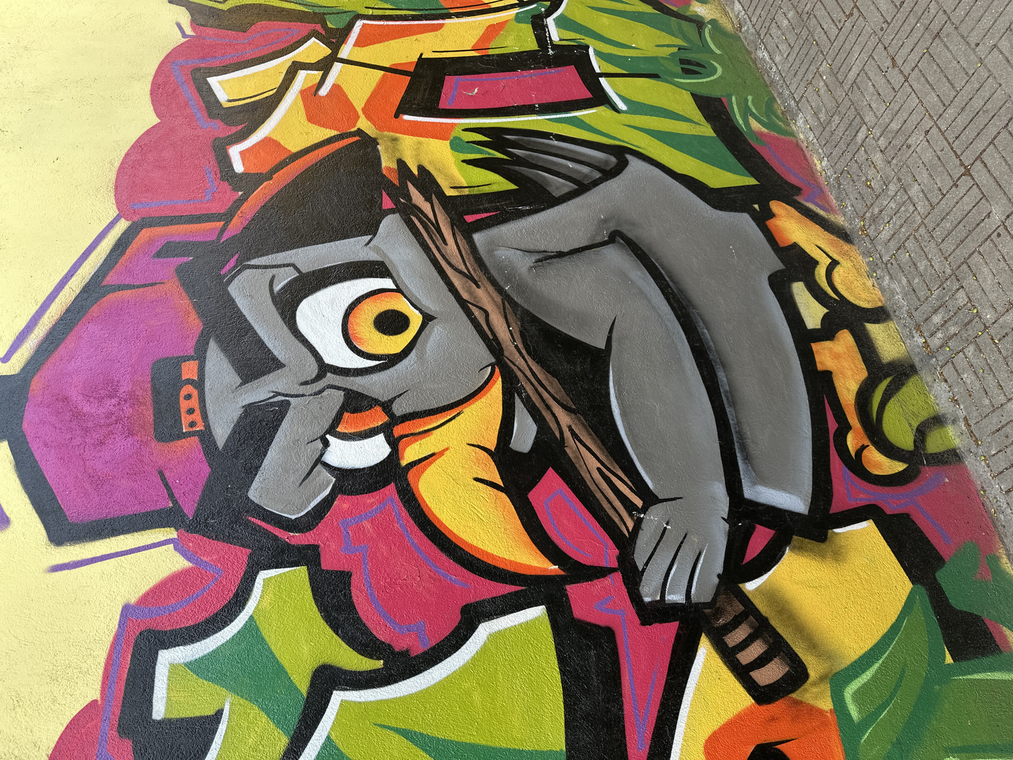 The image shows a section of graffiti art on a public surface, possibly a wall or pavement, located in Sofia, Bulgaria. The artwork features a cartoonish character with an exaggerated eye. The character is colored with shades of gray, yellow, white and red. It appears to be holding a wooden object, perhaps a stick or a weapon, rendered in brown tones. The background of the graffiti is a mix of vibrant colors including yellow, green, red, and purple. The graffiti is located on the edge of a paved area made of gray rectangular blocks.