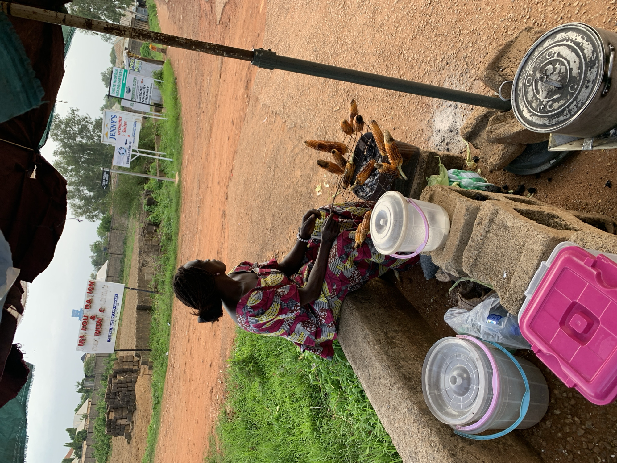 This image depicts an outdoor scene in Udu, Nigeria, likely a roadside stall. A woman, dressed in a vibrant, patterned fabric, sits on a stone ledge. In front of her, several ears of corn are arranged on a wire rack, suggesting they are being roasted or displayed for sale. Scattered around her are various plastic containers, including a large pink one and several buckets, some filled with water or other items. Behind the woman, a large metal pot with a lid sits on stones, possibly used for cooking or as part of the stall's setup.
The background reveals signs for local institutions, including "JENNY'S Apartments Suites," "TASTE NIGERIA HEADQUARTERS," "JOS INTERNATIONAL ACADEMY," and "IRIM FOUNDATION." These signs, along with the general earthy terrain and vegetation, provide context for the location. The lighting suggests it might be daytime, possibly overcast. The woman's posture and gaze, looking upwards, could imply a moment of rest or observation. The overall impression is of a typical day at a roadside vending spot in a Nigerian community.