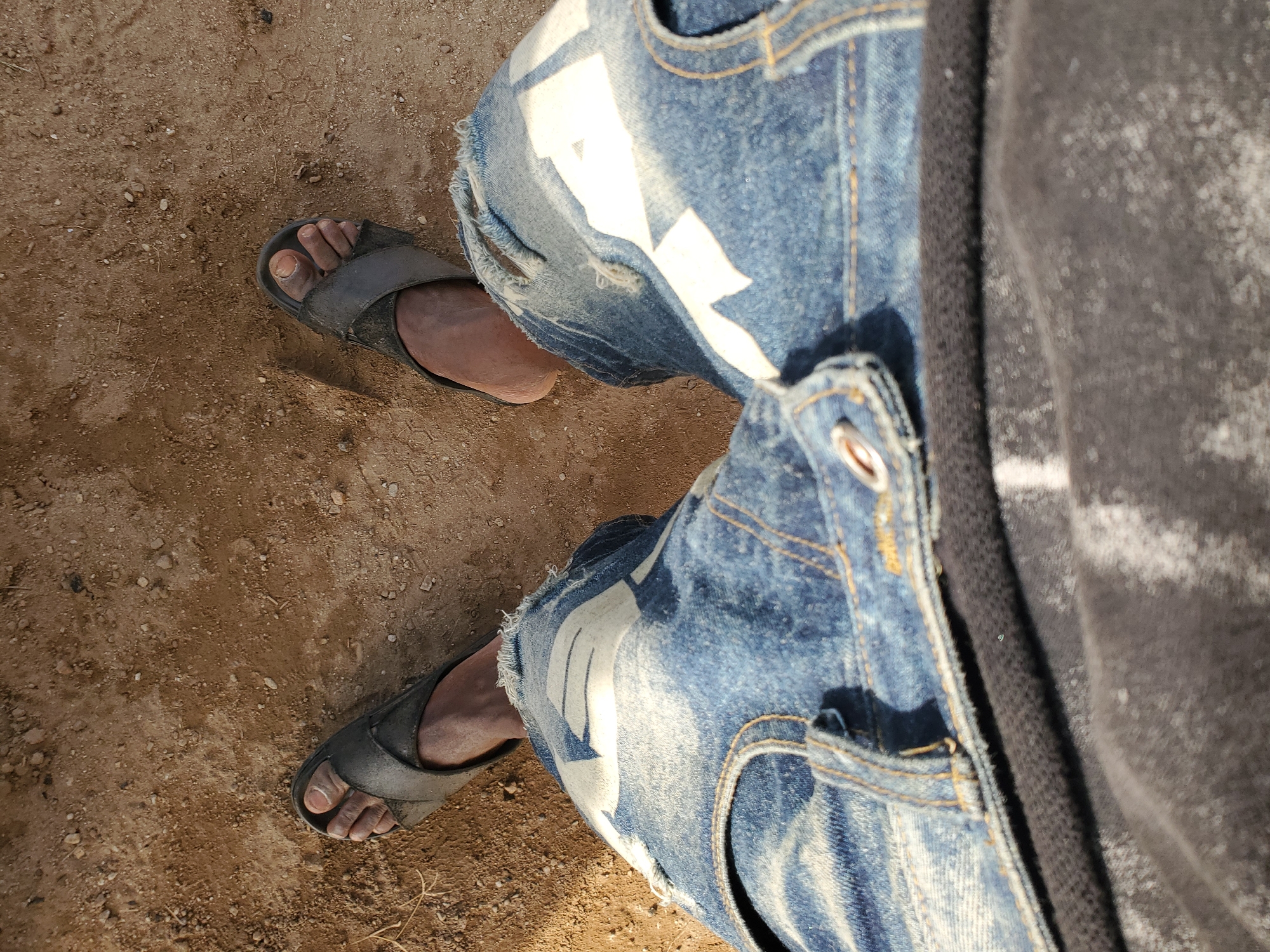 This image depicts a partial, top-down view of a person's lower body and feet, situated in Winters, United States of America. The individual is wearing distressed blue denim jeans with prominent faded white patterns on the thighs and frayed hems. A dark-colored garment, possibly a hoodie or jacket with a visible drawstring, is partially seen on the upper left. Both feet are shod in dark grey, open-toed sandals, resting on a dry, light brown ground composed of compacted earth, small pebbles, and scattered bits of dry vegetation. The exposed skin on the feet appears dark, and the toenails are visible, presenting a somewhat unkempt appearance. The scene is illuminated by bright daylight, casting subtle shadows.