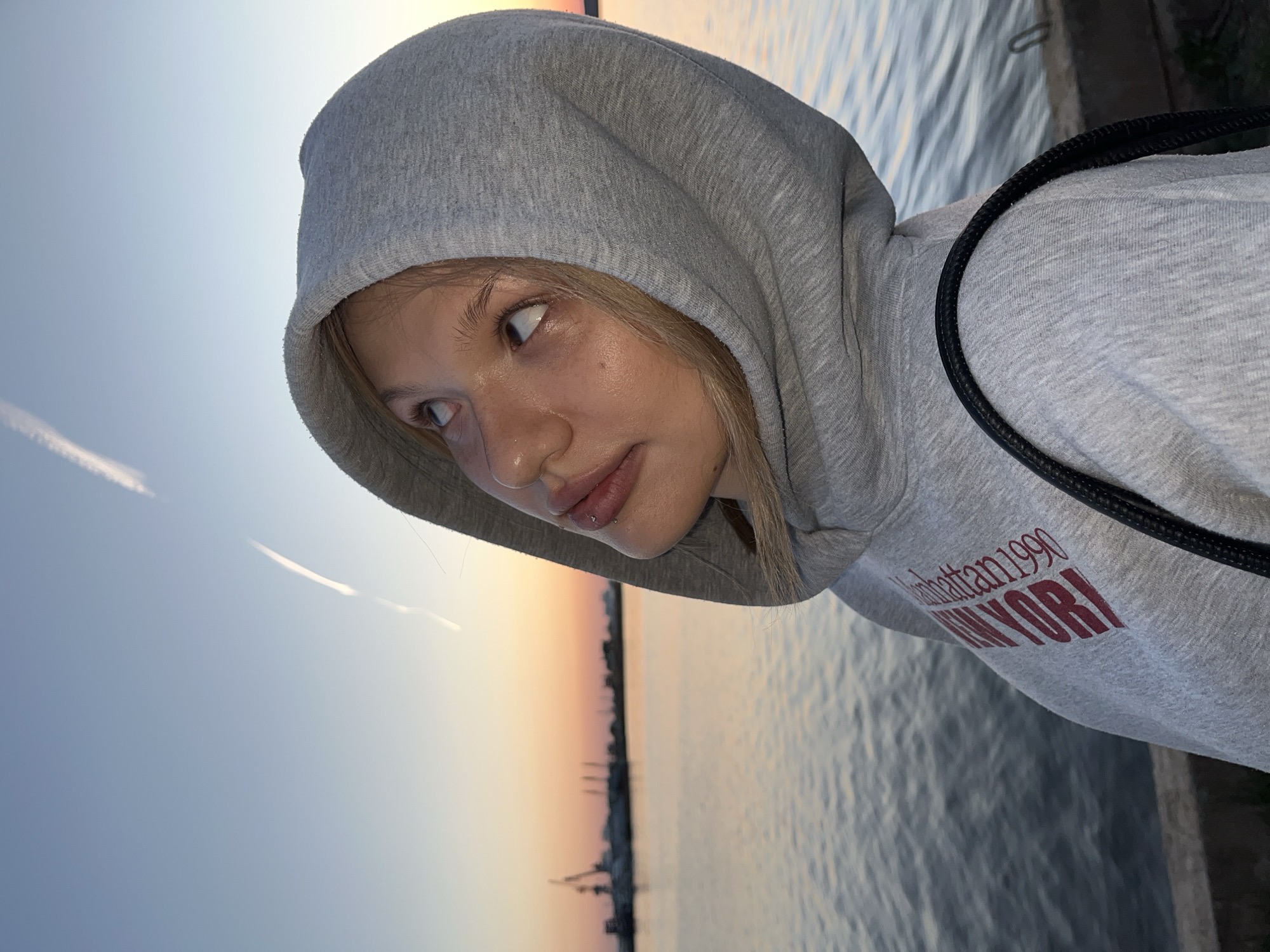 A person wearing a gray hoodie with the words "Manhattan 1990 NEW YORK" is looking off to the side. The background shows a body of water, possibly a river or bay, and a cityscape silhouetted against a sunset or sunrise. The image is taken at an angle, with the subject positioned on the right side of the frame. Based on the information provided, it is not possible to confirm if the location is in the city of Chaikovsky, Russia.