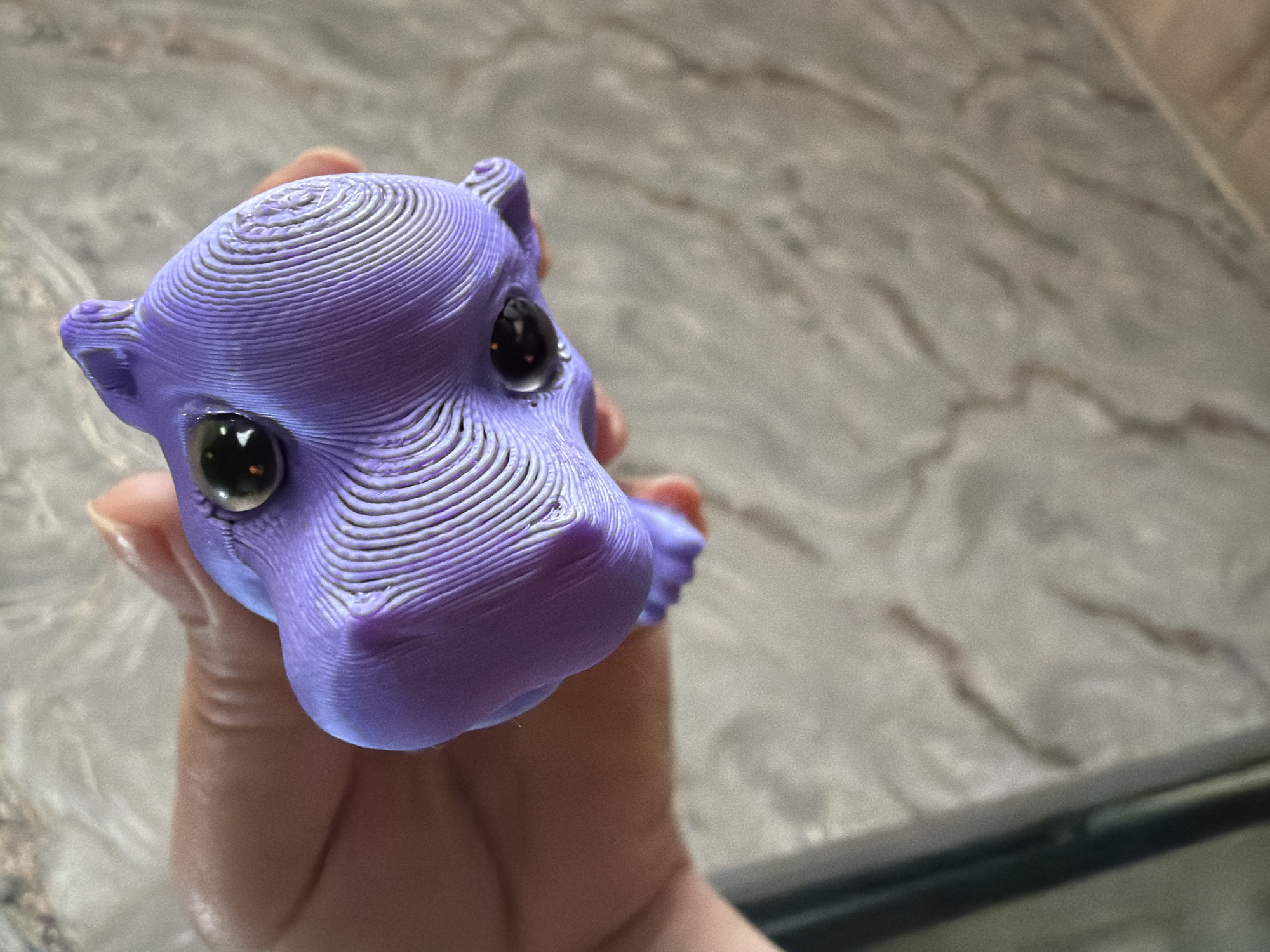 A close-up, slightly overhead shot shows a hand holding a purple 3D-printed toy with shiny black eyes. The toy is shaped like a cartoonish animal head, possibly a cat or a bunny, with visible layer lines from the printing process. The hand is positioned to support the toy from underneath. The background is a blurred, marbled surface, likely a countertop or table. The lighting is natural and diffused. There are no discernible landmarks or environmental cues to suggest a specific city or location.